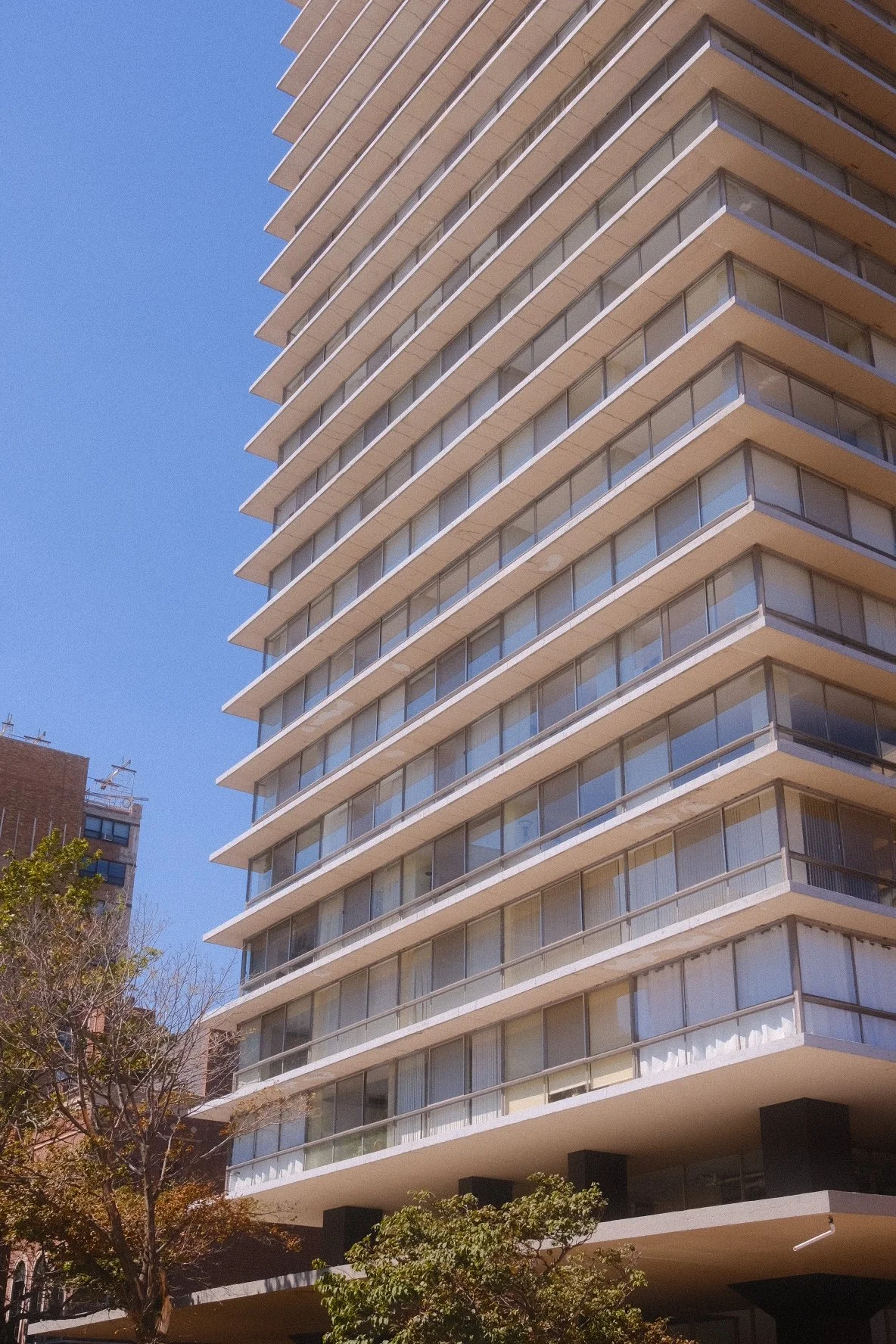 A tall midcentury-modern apartment building in Lakeview East Chicago with multiple balconies and glass windows, set against a clear blue sky, with some trees in the foreground.