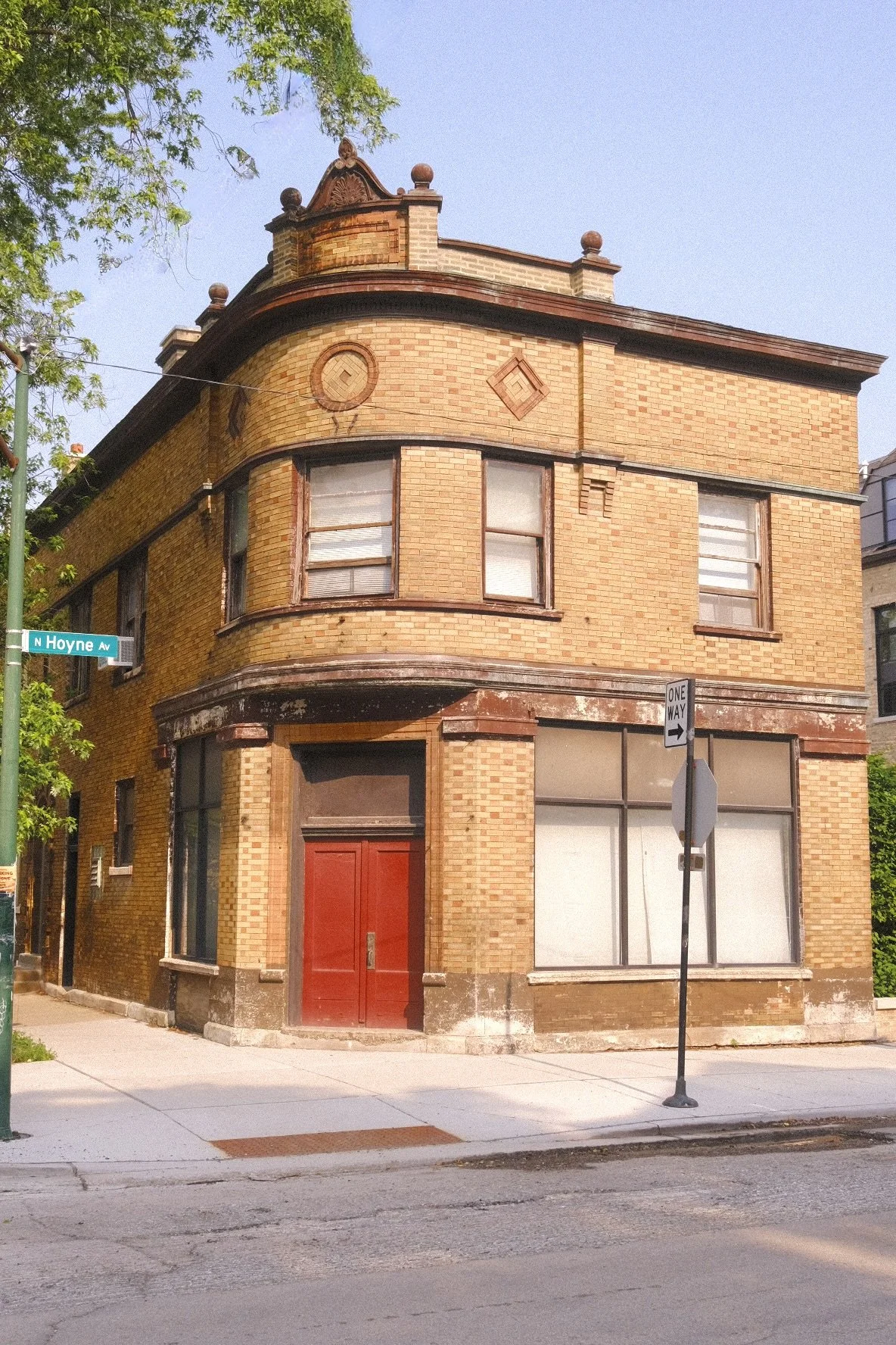 A three-story corner building in Bucktown made of yellow brick with large windows on all levels, a red door, and decorative architectural details at the top with some weathering and peeling paint at the bottom.