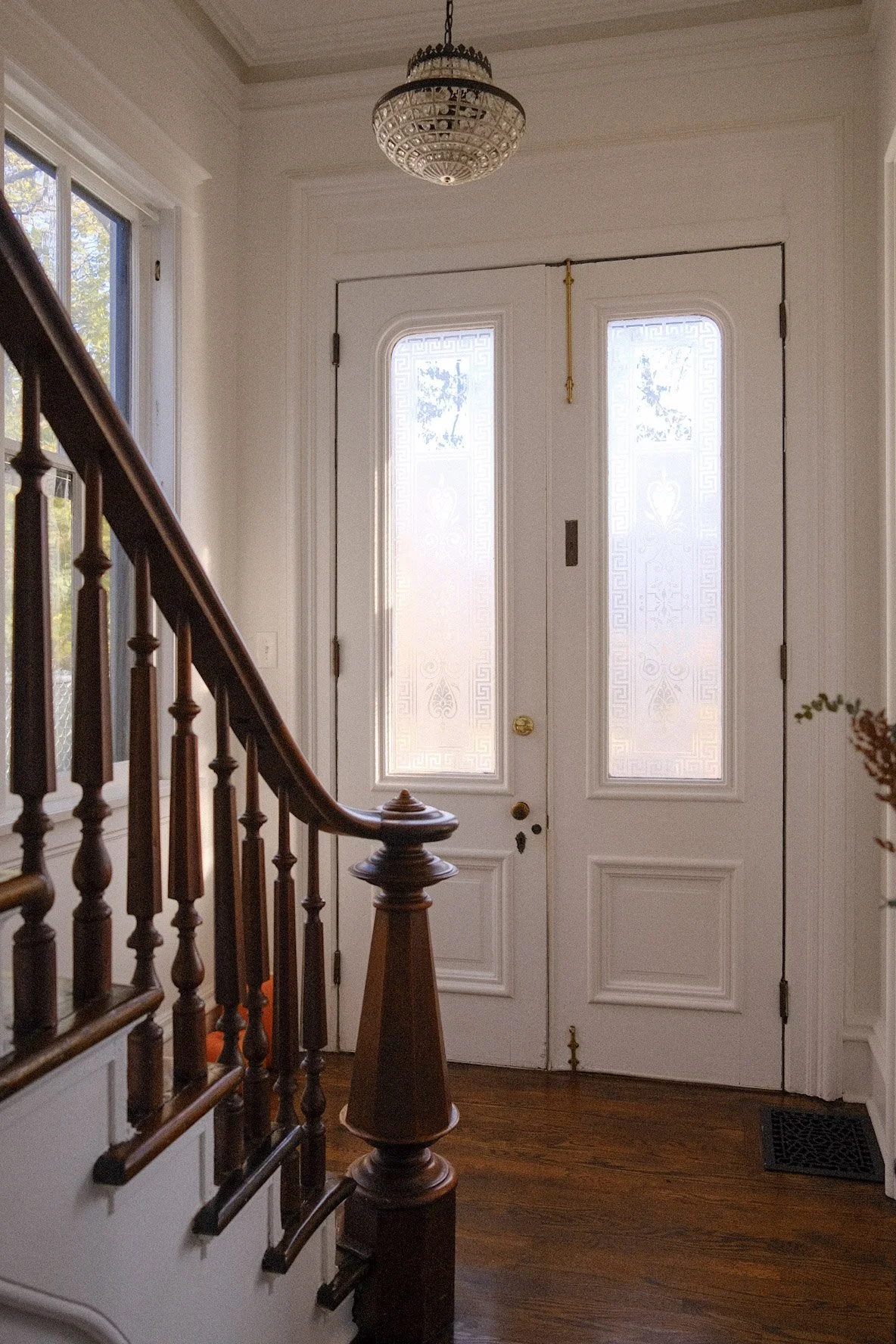 Interior view of an entryway featuring a white front door with decorative frosted glass panels, a brass doorknob, and a small brass lock. A wooden staircase with polished wood railing and spindles is on the left side, and there is a window with sheer