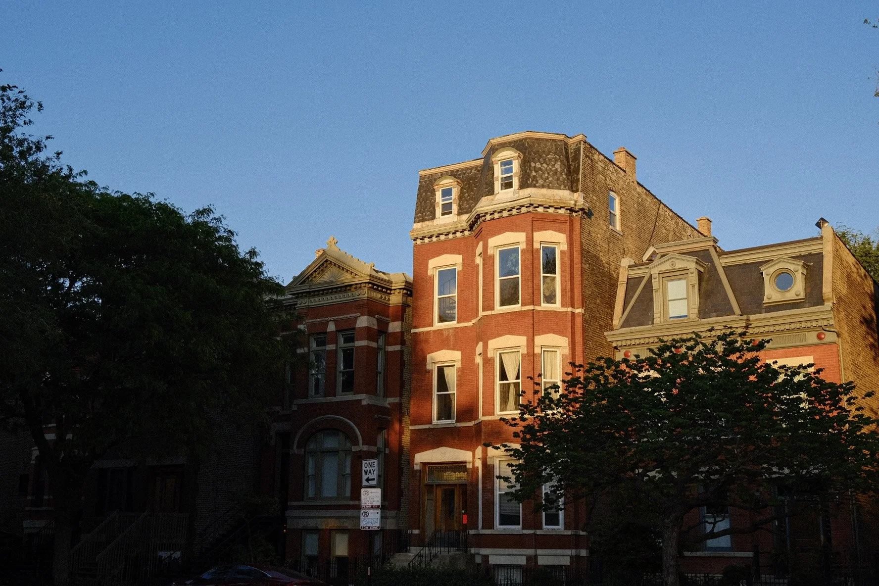 Sunset illuminating a historic red brick residential building with ornate architectural details, partially obscured by trees in an urban neighborhood.