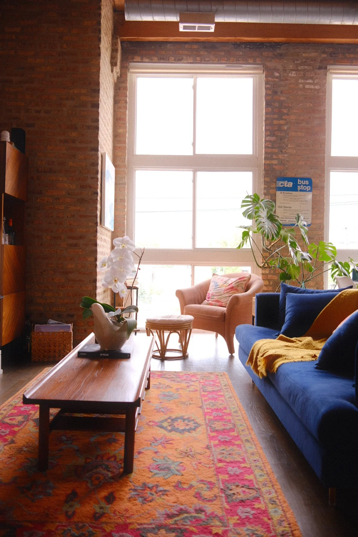 Living room with large windows, brick walls, a peach armchair with decorative pillow, dark blue sofa with yellow blanket, variety of plants including a large monstera, wooden coffee table with a vase of white orchids, colorful patterned rug, and a wo
