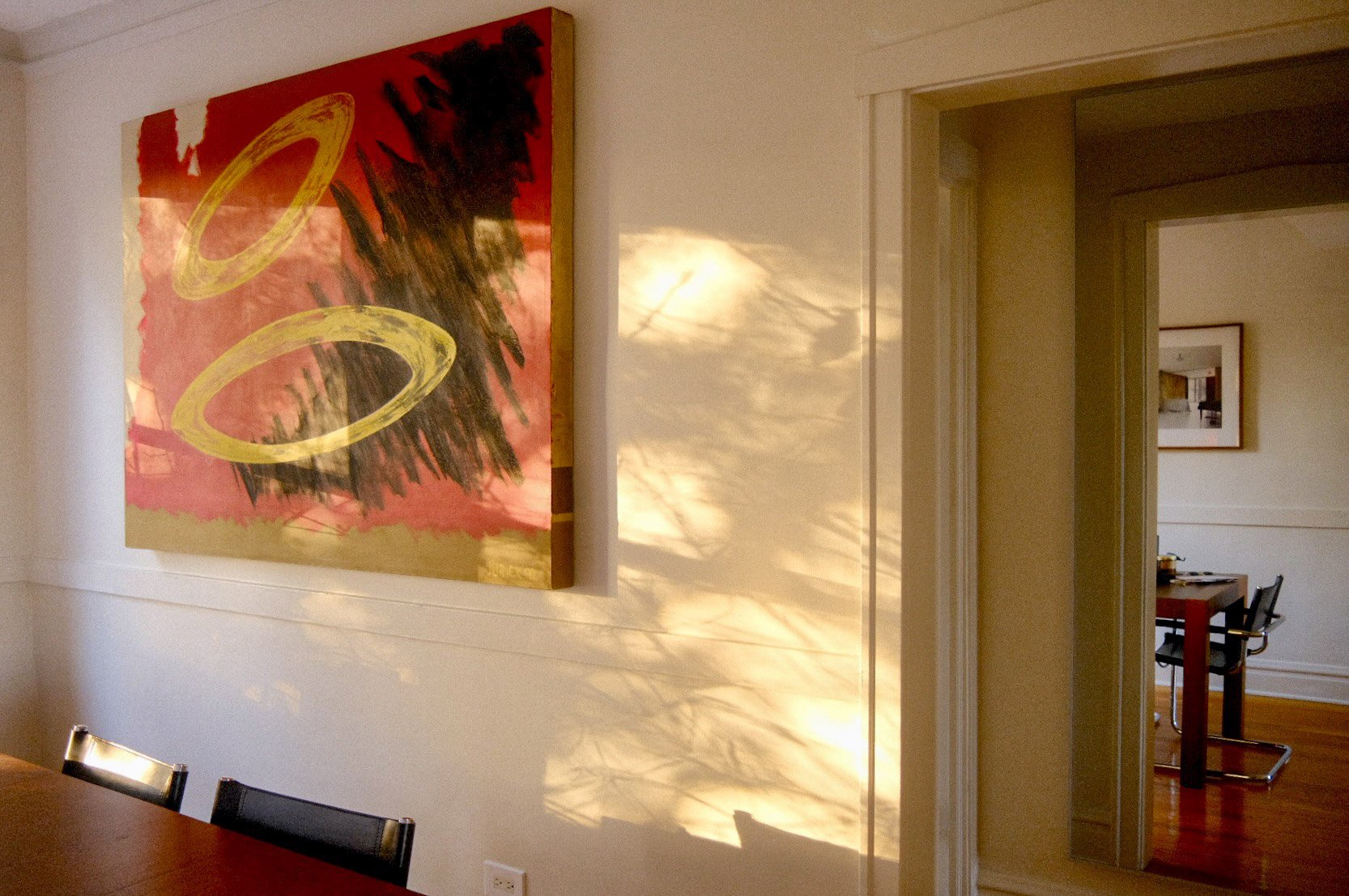 A colorful abstract painting with yellow oval shapes hanging on a white wall, with sunlight casting shadows on the wall and doorway leading to a room with a wooden table and chairs.