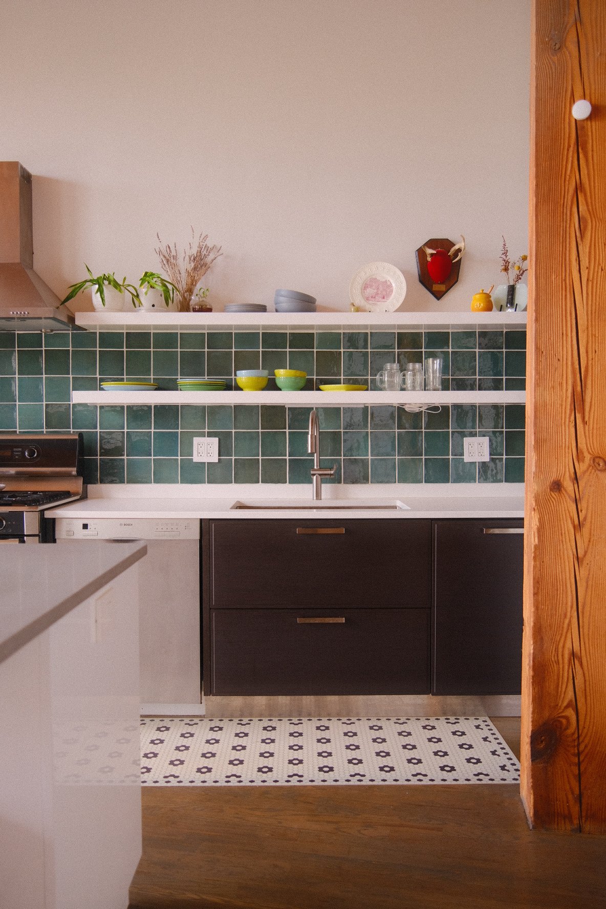 Kitchen with dark lower cabinets, green tiled backsplash, open shelves with dishes and plants, stainless steel sink, and decorative items on the wall.