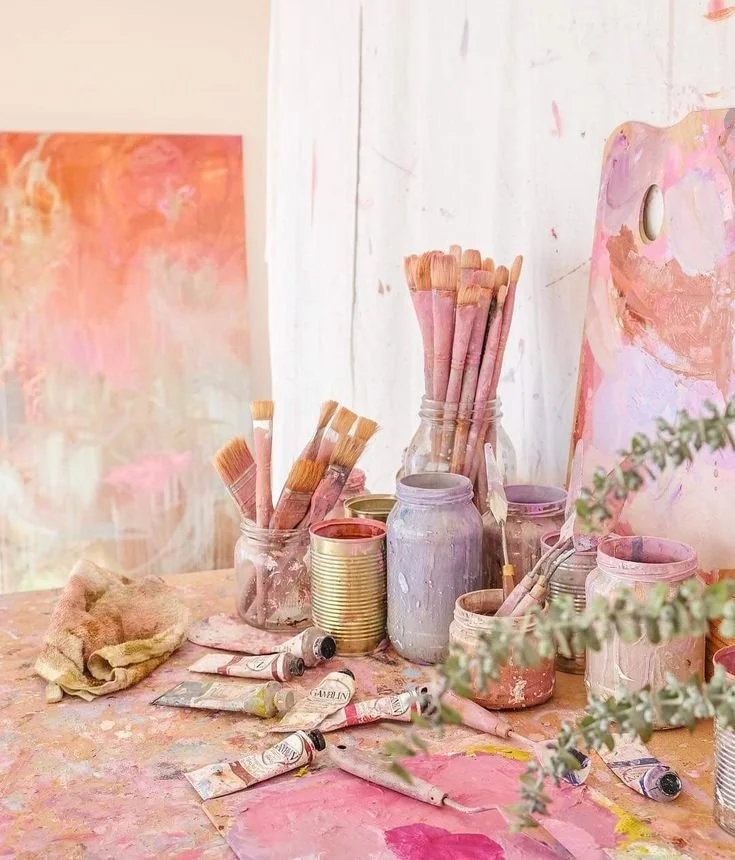 Art supplies, including brushes, paint tubes, and jars of paint, on a table with pink and pastel-colored artwork in the background.