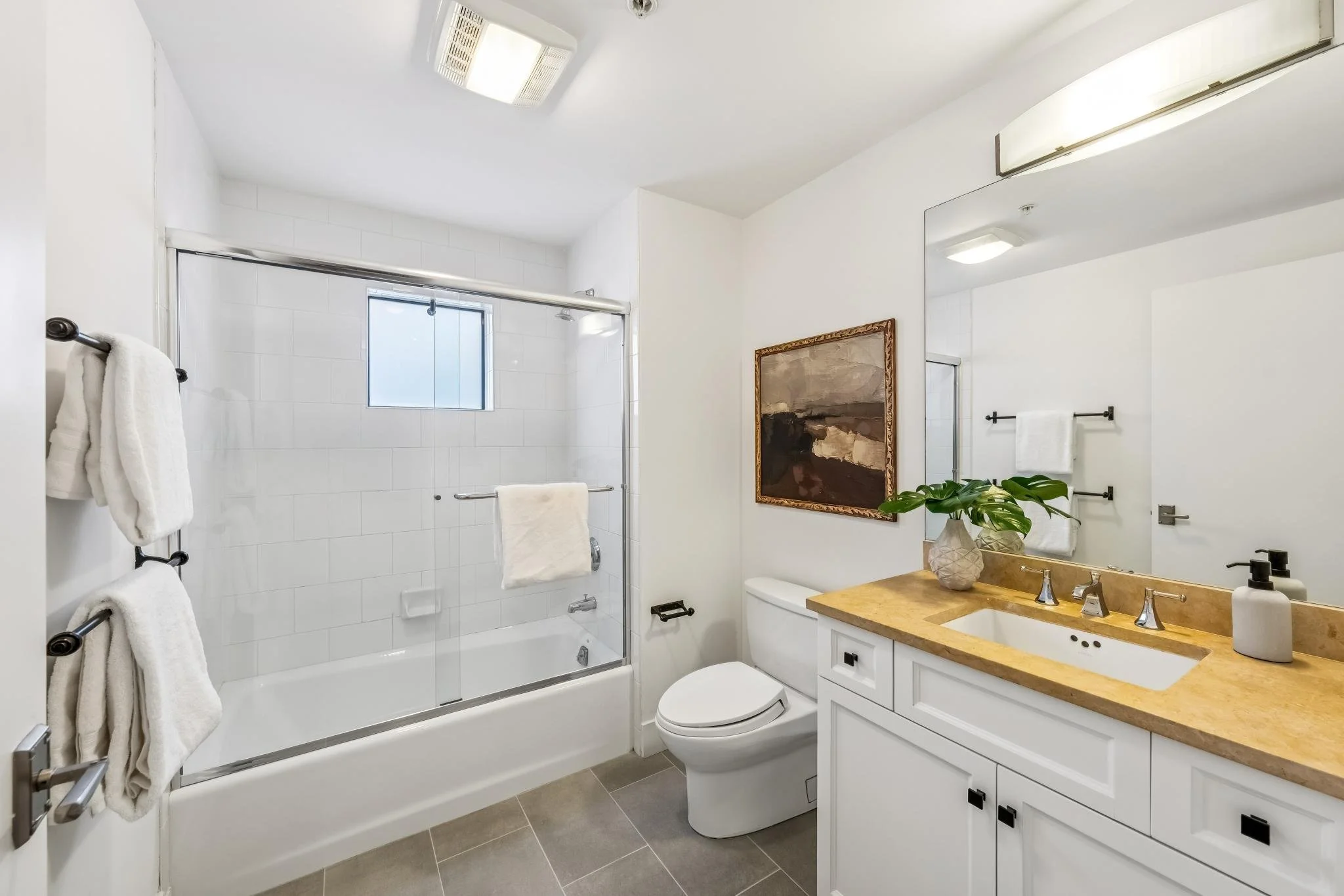 A modern bathroom with a glass-enclosed shower, white walls, a toilet, and a white vanity with a beige countertop. There are towels hanging on black towel bars, a framed painting on the wall, and a potted plant on the vanity. The bathroom has a ceili