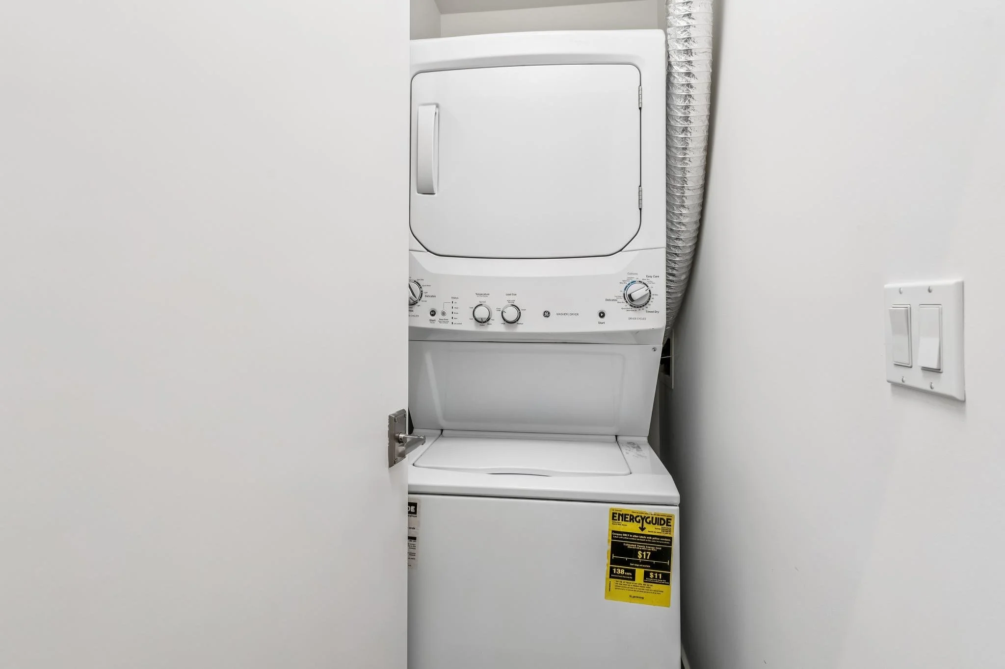 Front view of a white stacked washer and dryer in a laundry area, with a silver vent pipe on the right and an energy guide label on the lower right corner.