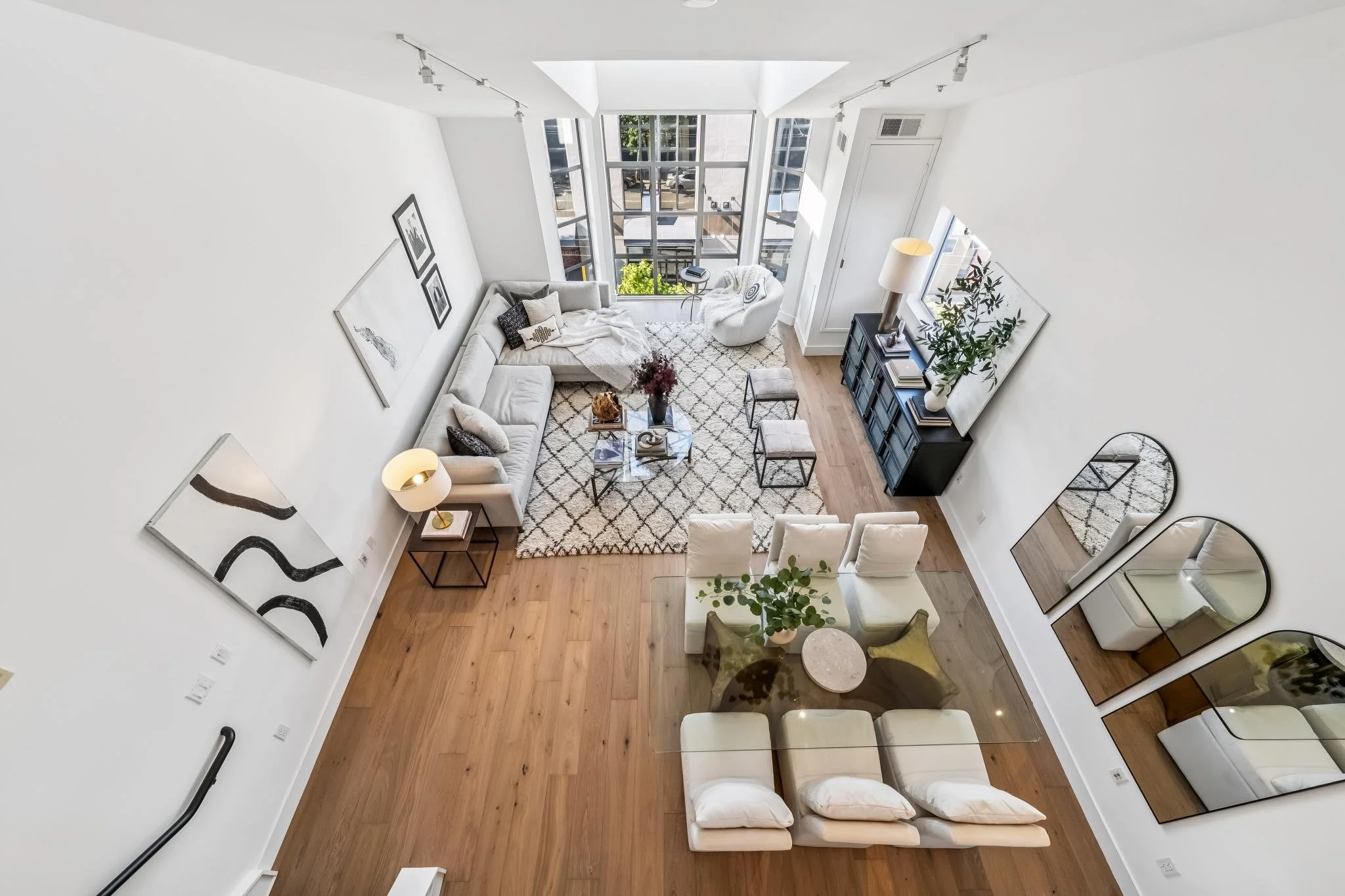 A spacious, modern living room with white walls, a beige sectional sofa with black and white pillows, a white armchair, a wooden coffee table, a black TV stand with books, and large windows bringing in natural light. The room has a wooden floor with 