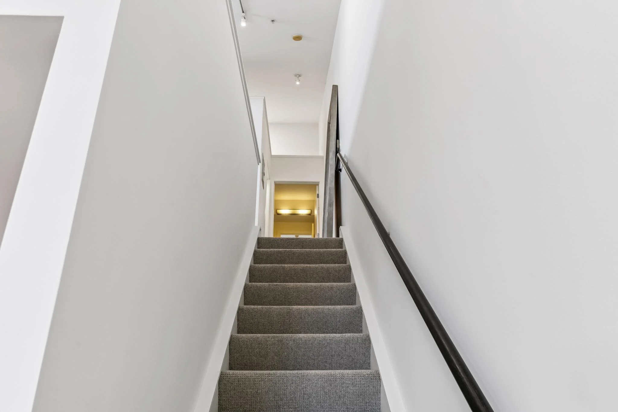 Indoor staircase with carpeted steps and a dark handrail on the right, leading up to a hallway with a doorway and yellow lighting at the end, white walls, and a ceiling with lights.