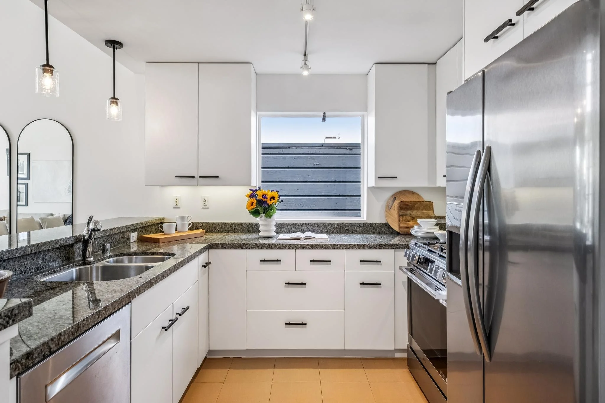 Modern kitchen with white cabinets, gray granite countertop, stainless steel appliances, a window with a view of the sky and a wooden fence, a vase with yellow and purple flowers, a cutting board, dishes, and hanging light fixtures.