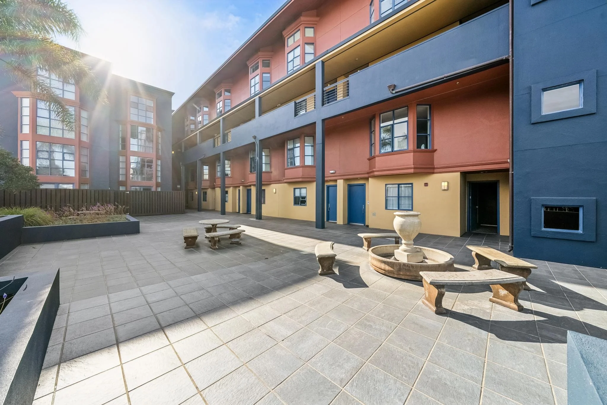 Modern multi-story apartment building with colorful facade including blue, yellow, and reddish sections. There is a courtyard with outdoor seating, planting beds, and a fountain, illuminated by sunlight.