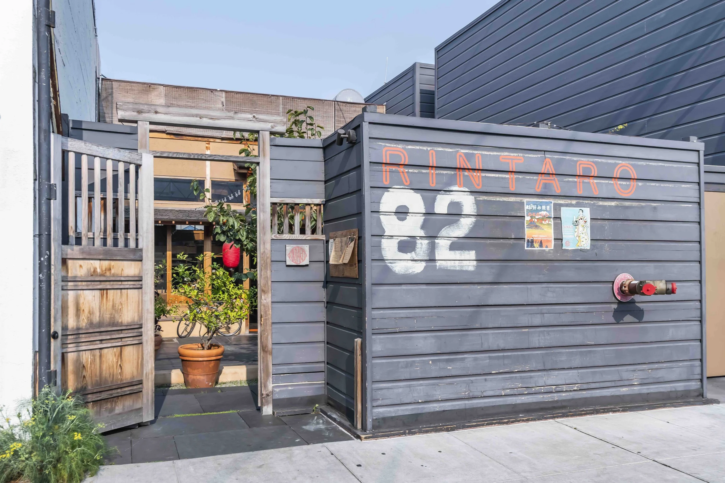 A modern outdoor space with a gray wooden fence and a sign reading 'RINTARO' in orange neon lights. Inside the space, there are plants, lanterns, and a small bicycle. There is also a small structure made of gray wood with a fire hydrant attached to i
