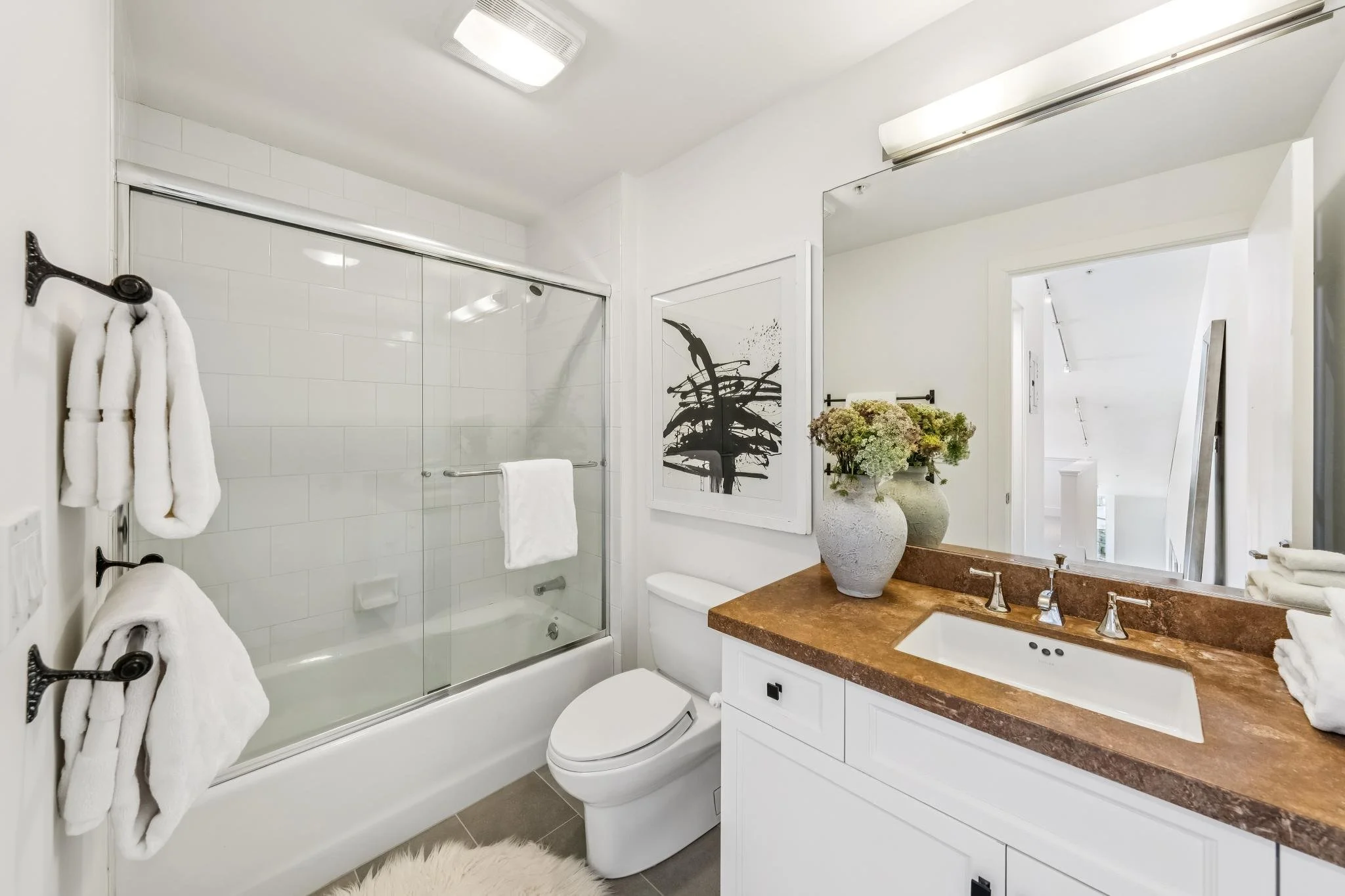 A modern bathroom with white tiles, a glass shower enclosure, a white toilet, a wooden vanity with a brown countertop, a large mirror, and white towels on black towel racks. There is a flower arrangement on the countertop and a framed art piece on th