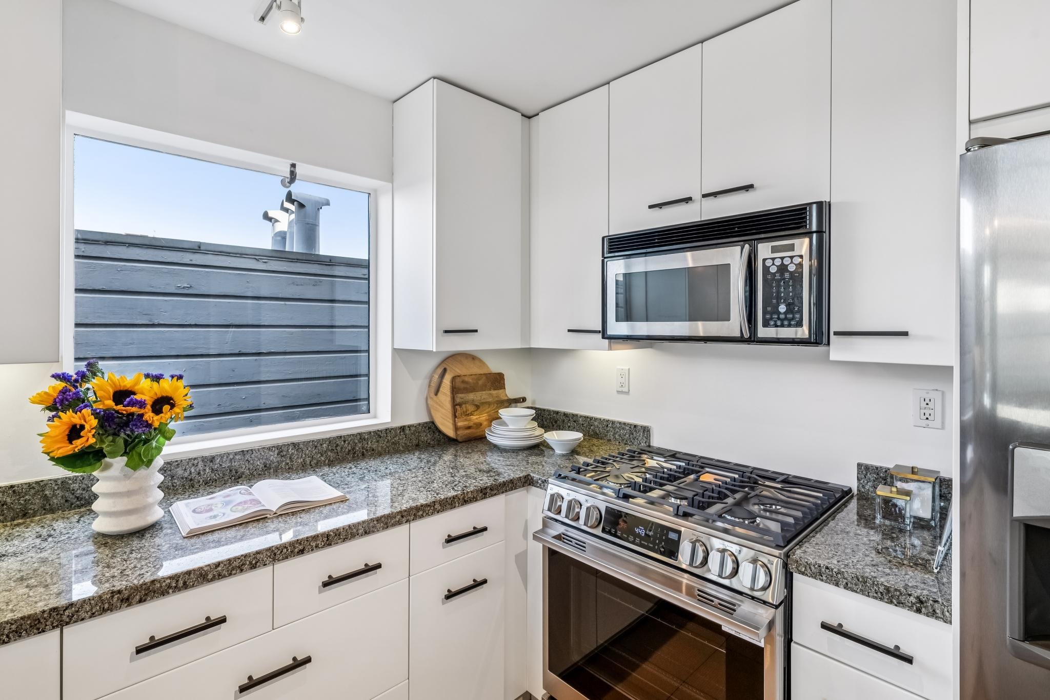A modern kitchen with white cabinets, granite countertop, and stainless steel appliances including a microwave and stove. There is a window with a view of a gray wooden fence outside, a bouquet of sunflowers and purple flowers on the counter, an open