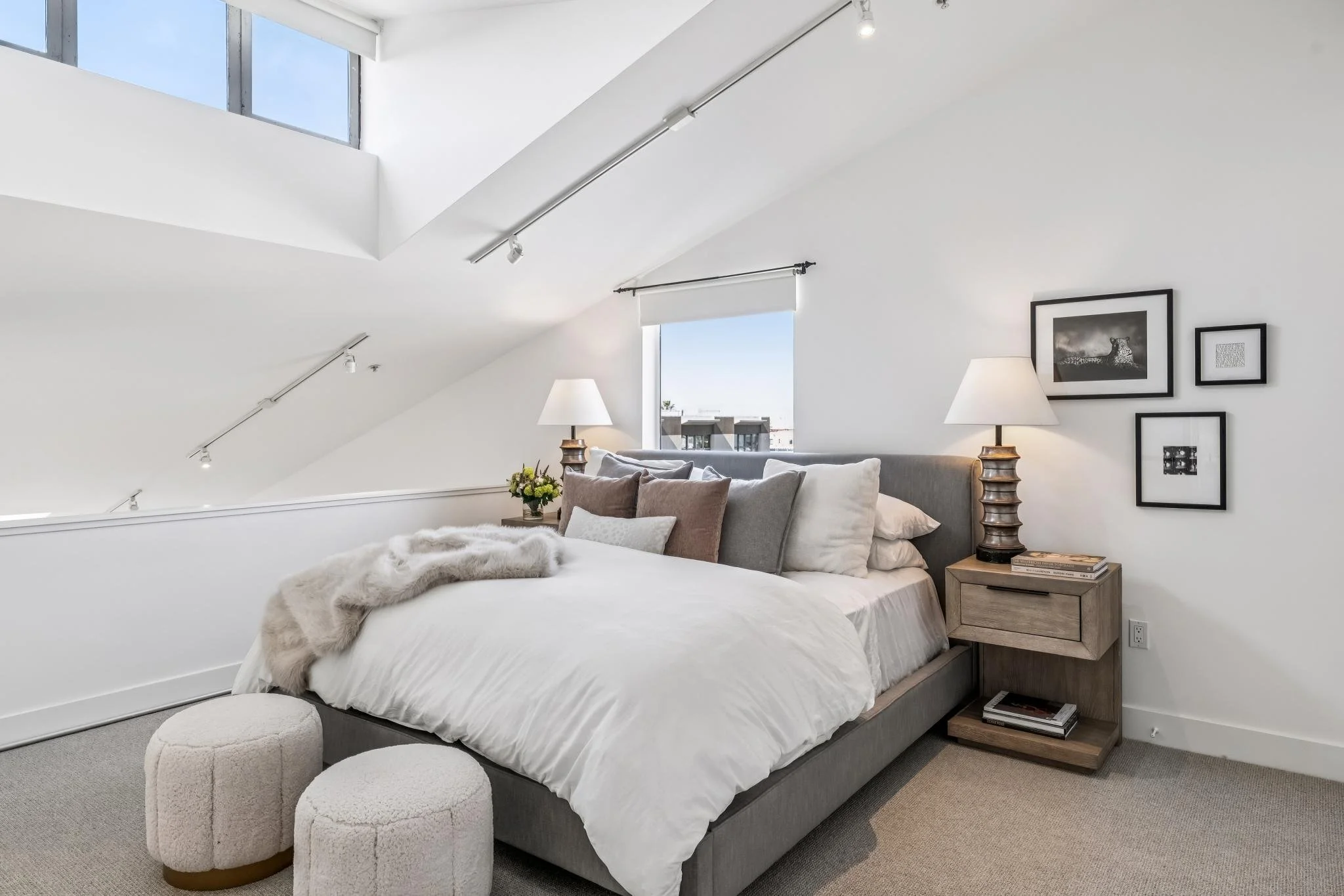 A modern bedroom with a sloped ceiling, a skylight window with blue sky, a bed with white linens and pillows, two small white ottomans at the foot of the bed, two white lamps on bedside tables, a window with white blinds, and framed black and white a