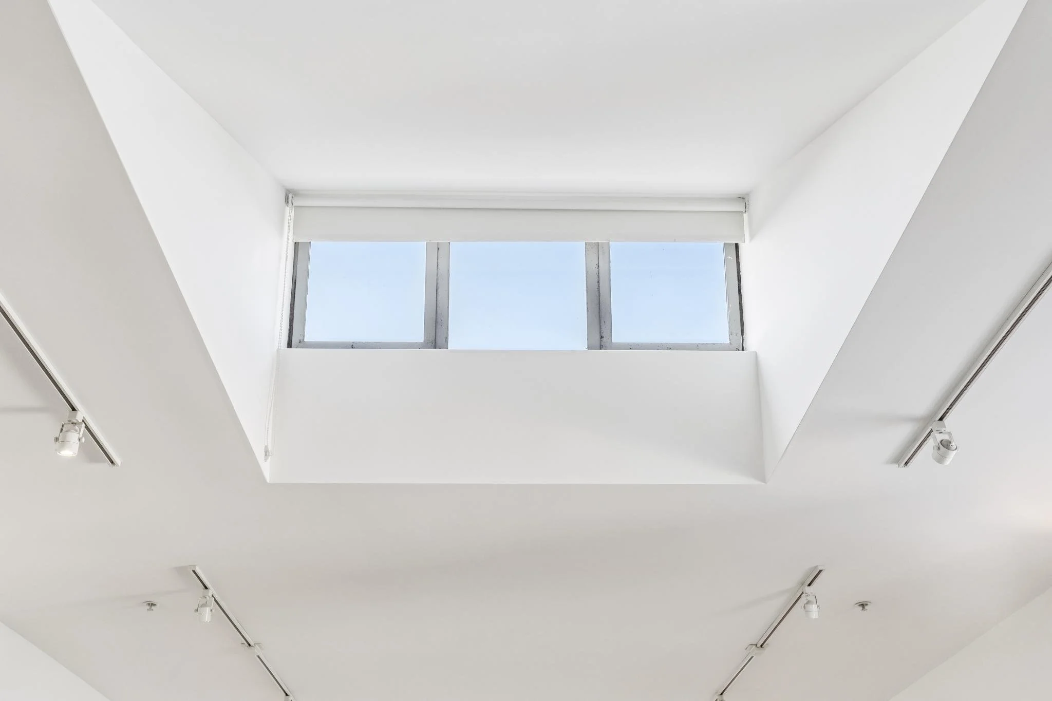 A view of a white ceiling with a skylight window showing a clear blue sky, surrounded by white walls and track lighting fixtures.