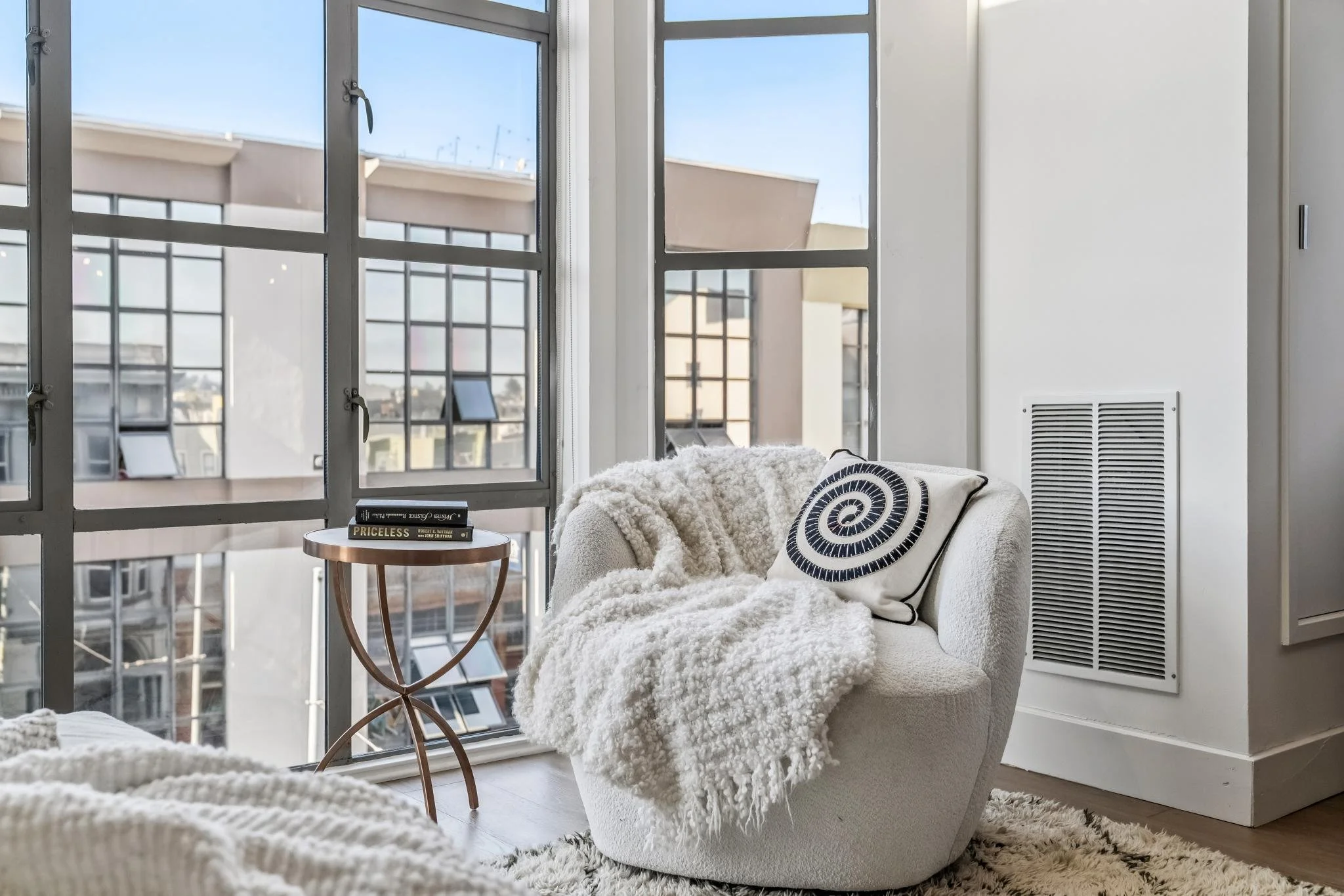 A cozy white armchair with a spiral patterned pillow, a blanket, and a round side table holding books in front of large windows with grid panes, overlooking modern buildings and a blue sky.