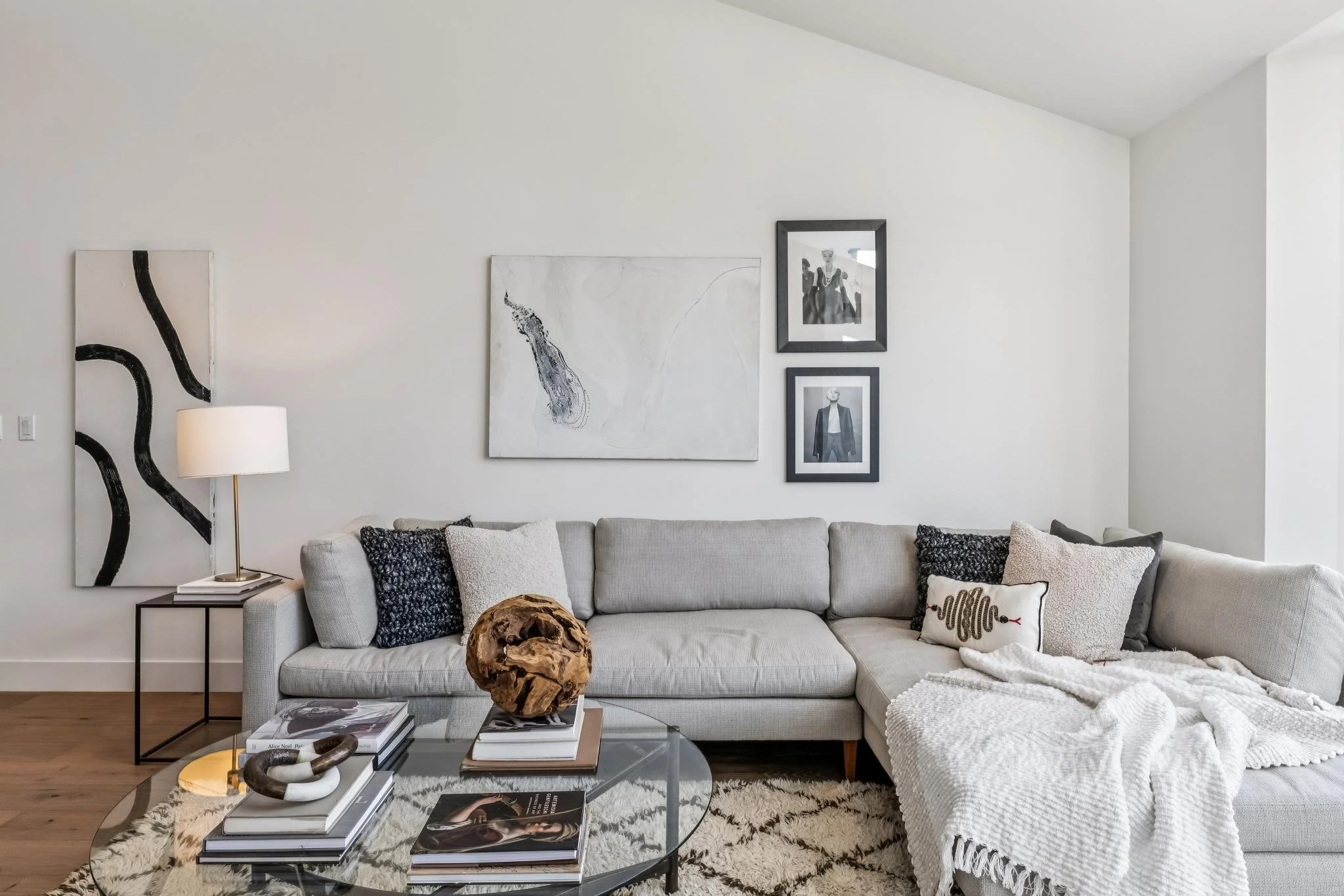 A modern living room with a white sofa, black and white throw pillows, a glass coffee table with books and a decorative wooden sculpture, a white wall with black and white framed art, a black side table with a white lamp, and a beige and brown area r