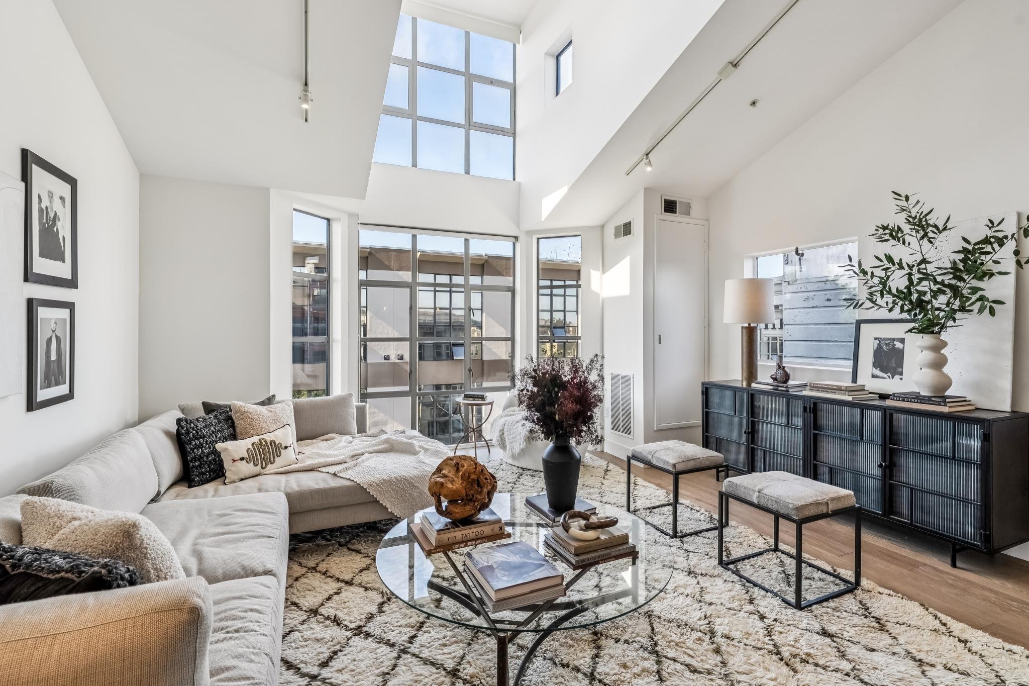 Bright modern living room with large windows and skylight, white walls, beige sectional sofa with decorative pillows, glass coffee table with books and decorative objects, black console with framed photos and potted plant, and a patterned rug.