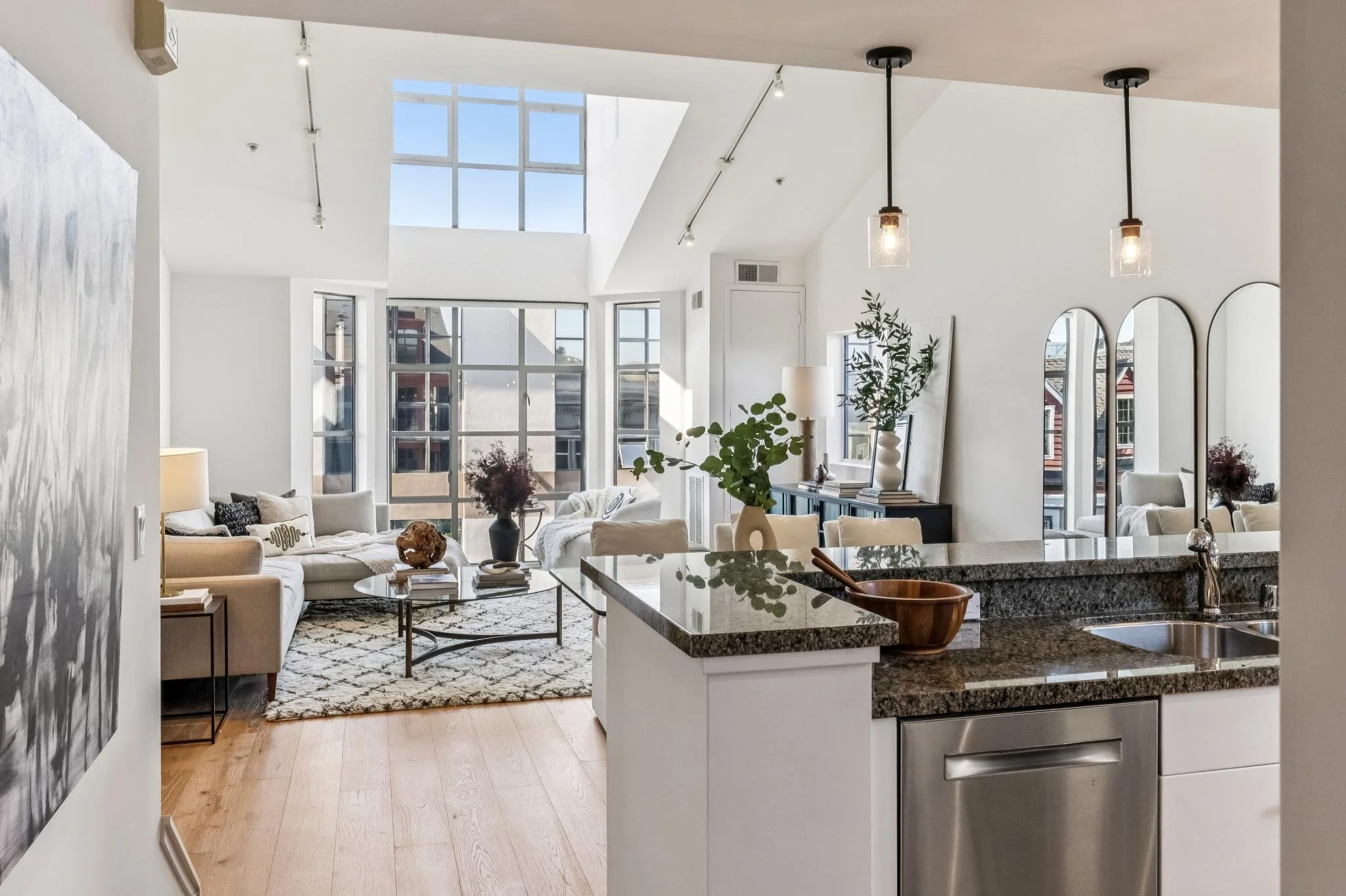 Modern living room with large windows, white walls, and contemporary furniture, seen from the kitchen area with granite countertops.
