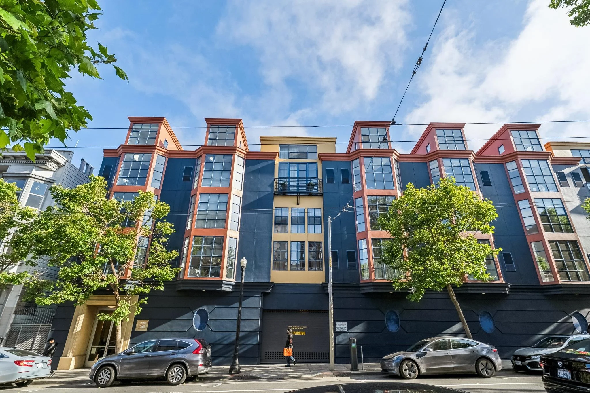 Modern multi-story residential building with large glass windows, colorful accents, and a small balcony. Trees line the street in front, with parked cars and pedestrians visible.