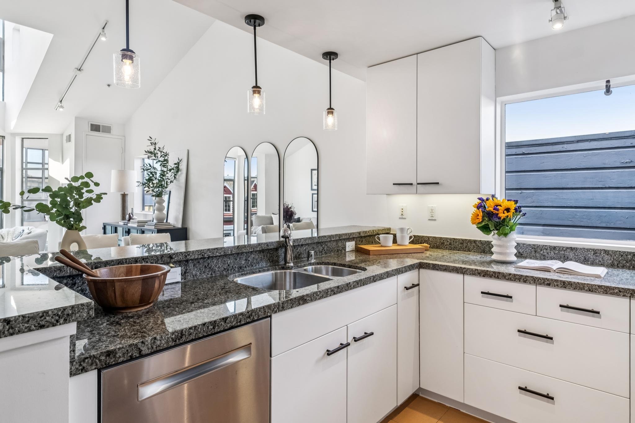 Modern kitchen with white cabinets, black handles, granite countertops, and a double sink. There are hanging pendant lights, a vase with sunflowers on the countertop, potted plants, and a window with blue sky visible outside.