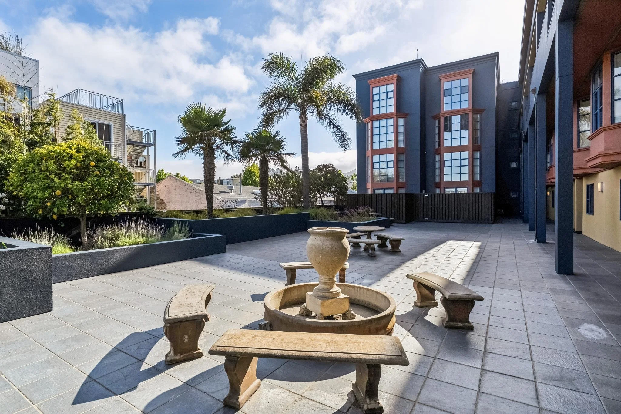 A courtyard with a tile floor, surrounded by modern apartment buildings, featuring trees, benches, and a central stone fountain.