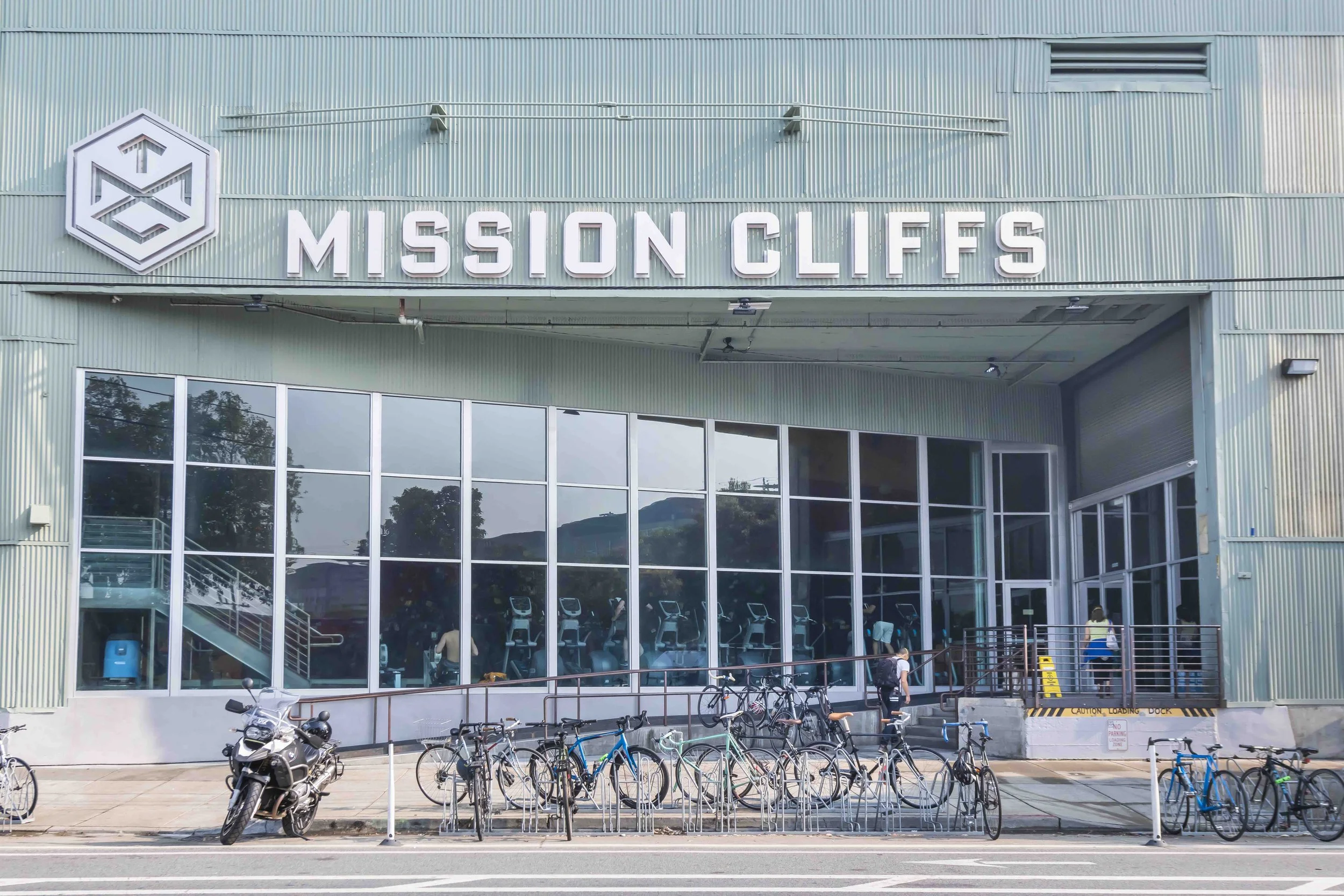The exterior of a gym named Mission Cliffs with bicycle racks and a motorcycle in front, large windows revealing indoor exercise equipment, and a sign displaying the gym's name.