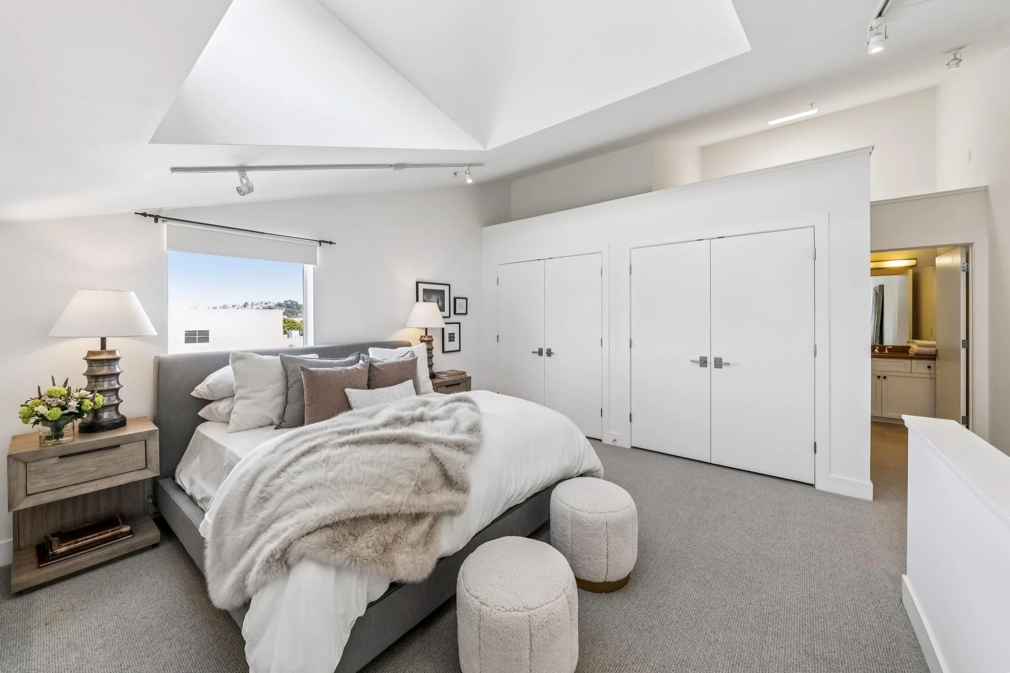 A modern, minimalist bedroom with white walls and ceiling, gray furniture, and a large bed with white and beige bedding. There are two small, round, white stools in front of the bed. On the bedside table, there are two lamps with beige shades and a b
