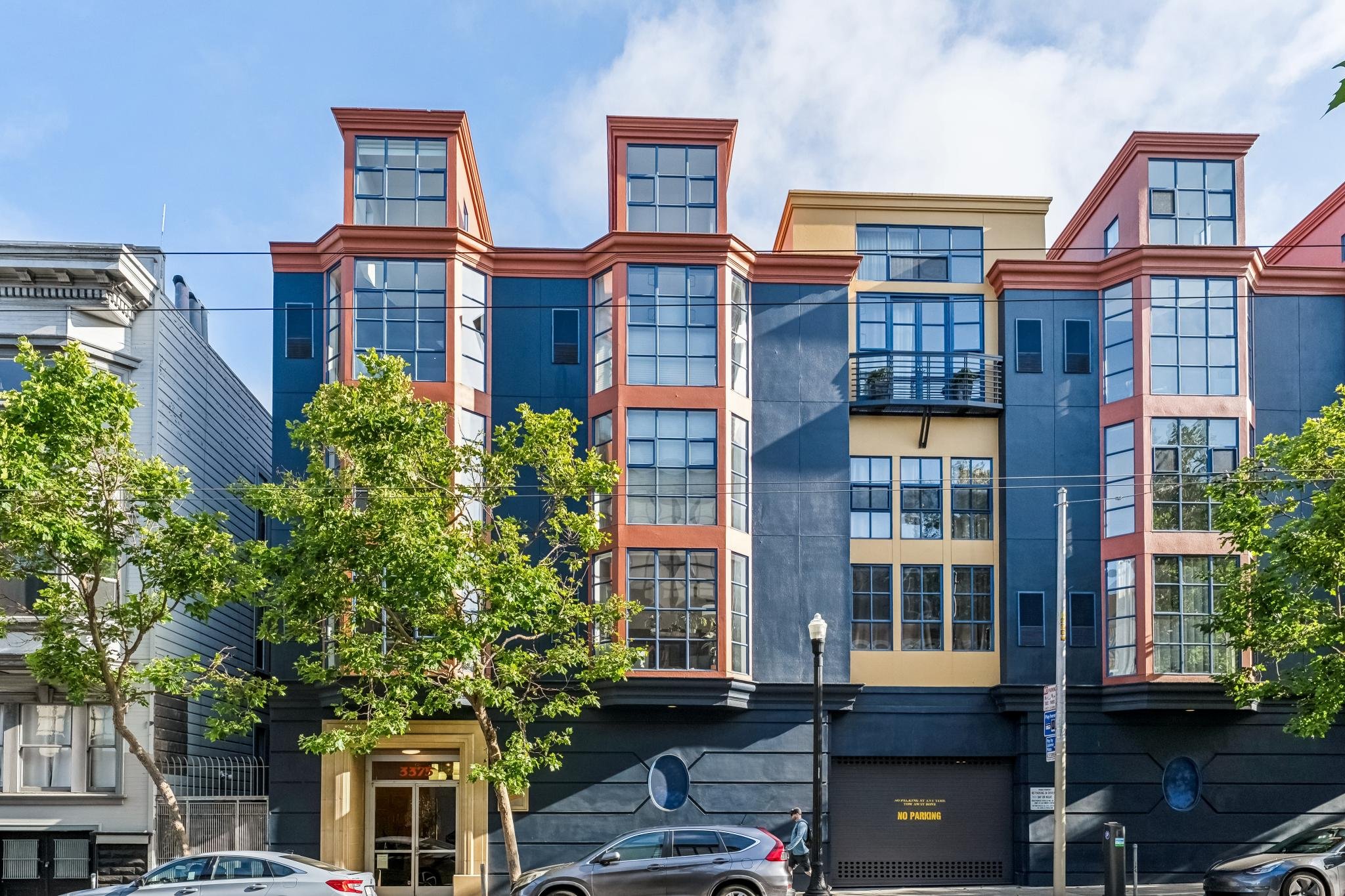 Colorful modern apartment building with large windows and a blue, yellow, and pink facade, surrounded by green trees and parked cars, under a partly cloudy sky.