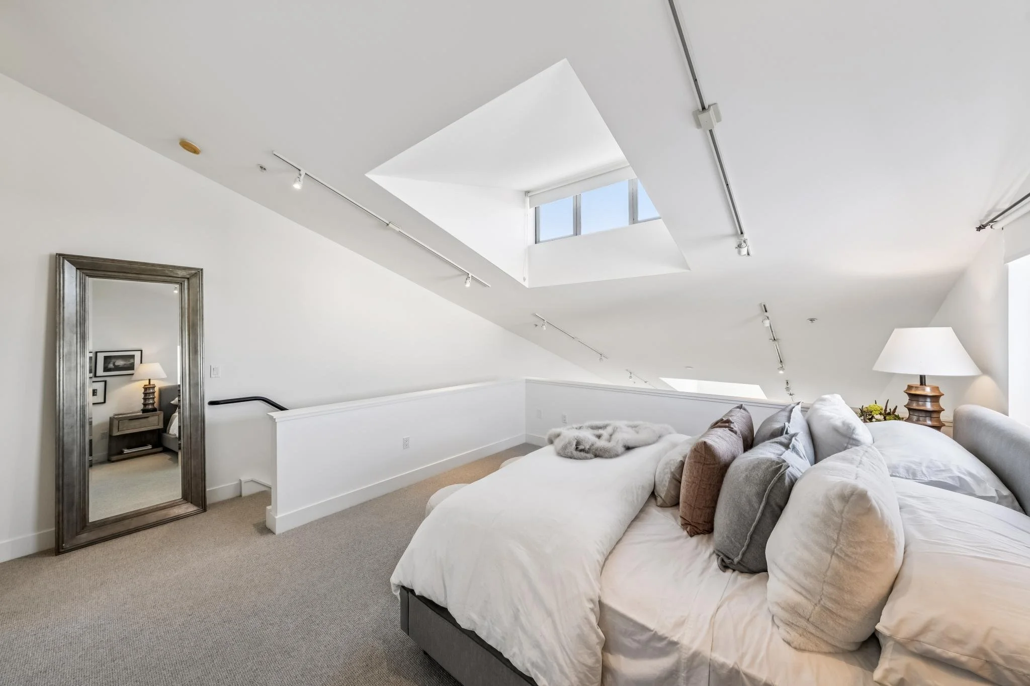 A modern bedroom with white walls, a large bed, beige carpet, and a skylight ceiling with natural light. There is a mirror leaning against one wall, a small nightstand with a lamp, and a pile of pillows and a fuzzy blanket on the bed.