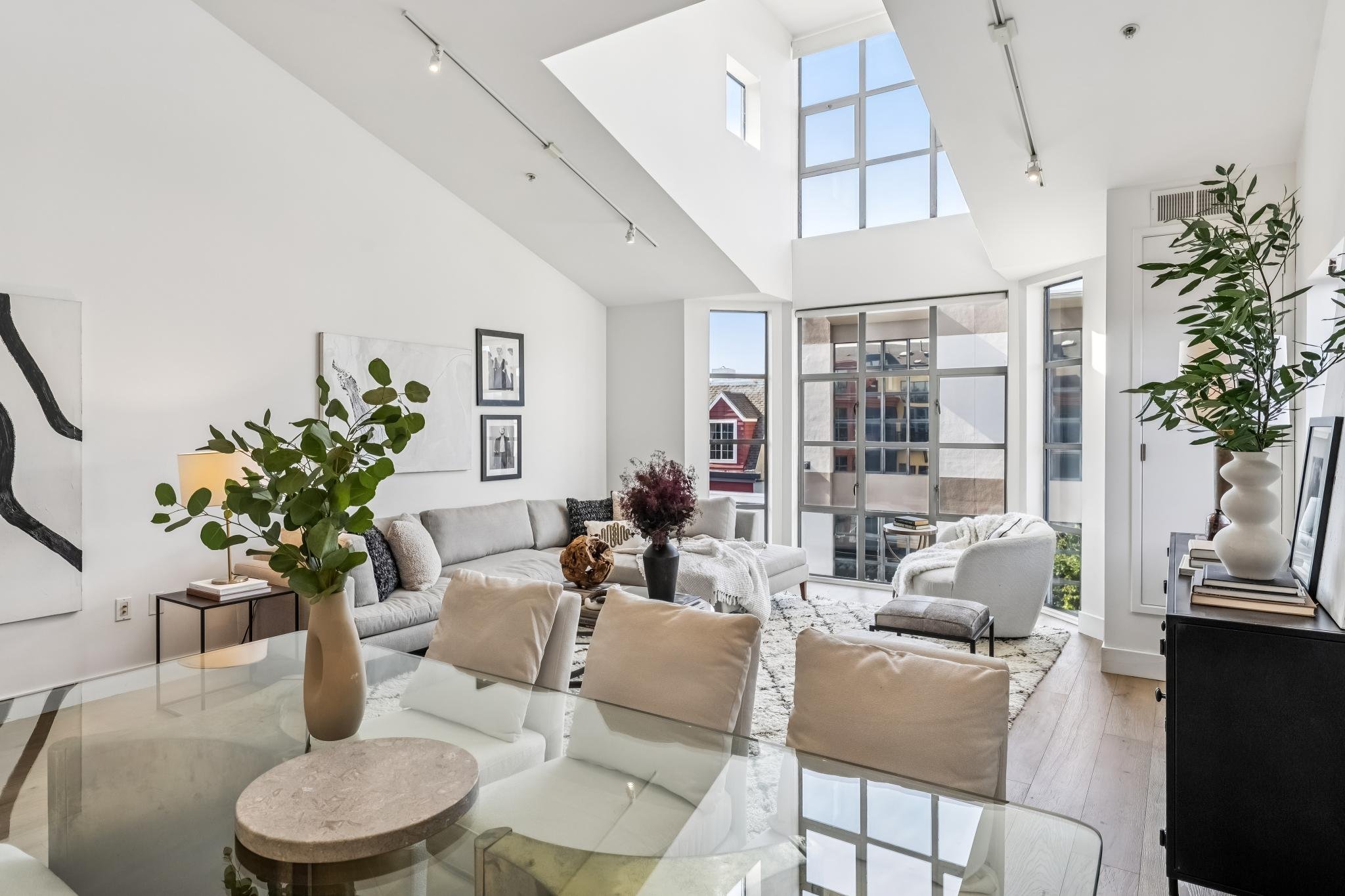 Modern living room with white walls, large windows, and high ceiling. Contains a beige sofa, white armchair, glass coffee table with a stone centerpiece, and decorative plants. Black and white artwork on the wall, and sunlight streaming through windo