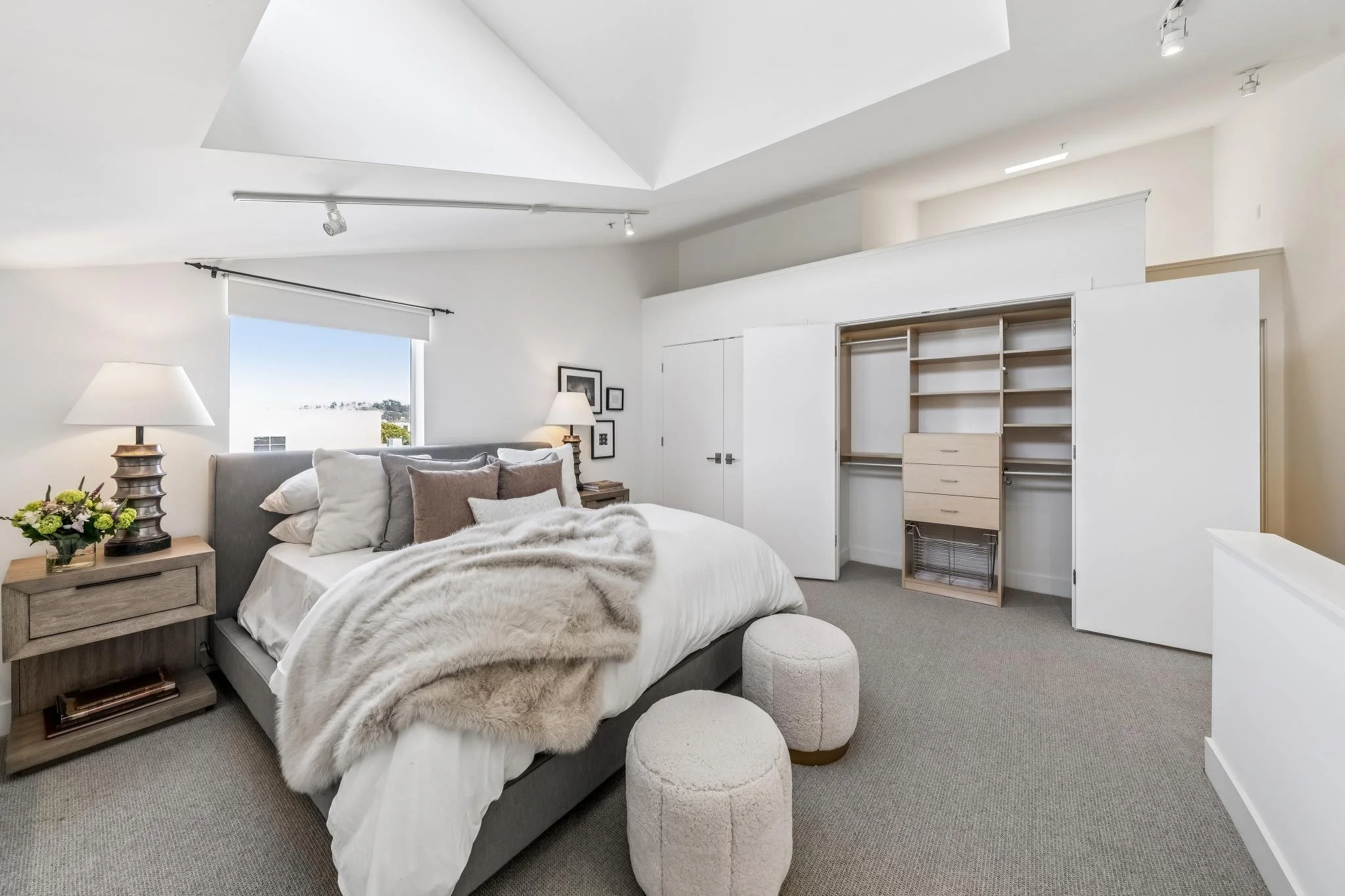 A bedroom with a large bed, nightstand with a lamp, plant, and books, a window with a view outside, and a built-in open closet with drawers and hanging space, decorated in neutral tones and soft lighting.
