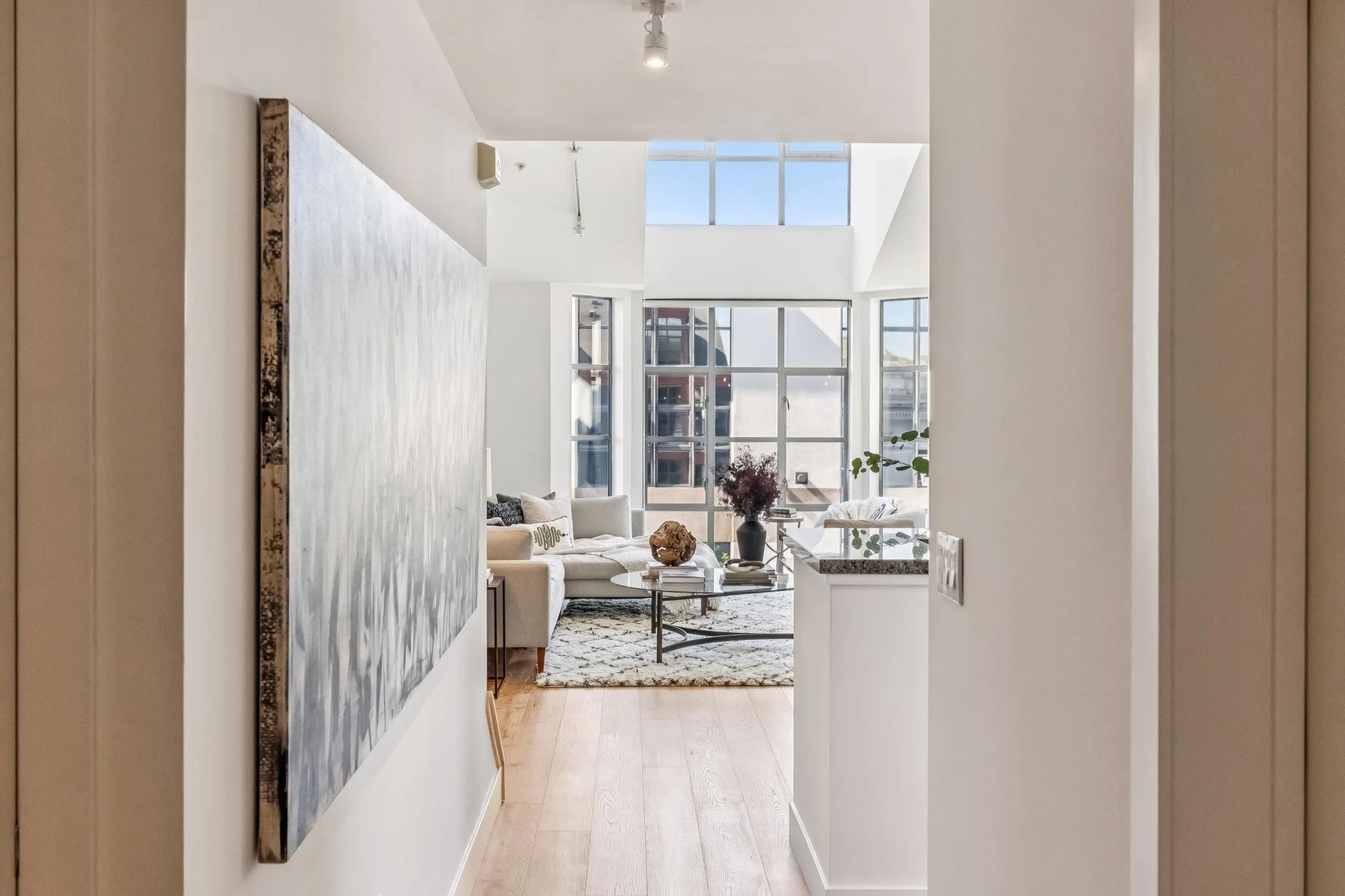 View through a hallway into a bright, modern living room with large windows, white walls, a white sofa with cushions, a glass coffee table, a patterned rug, and decorative items including a large vase with flowers and books.