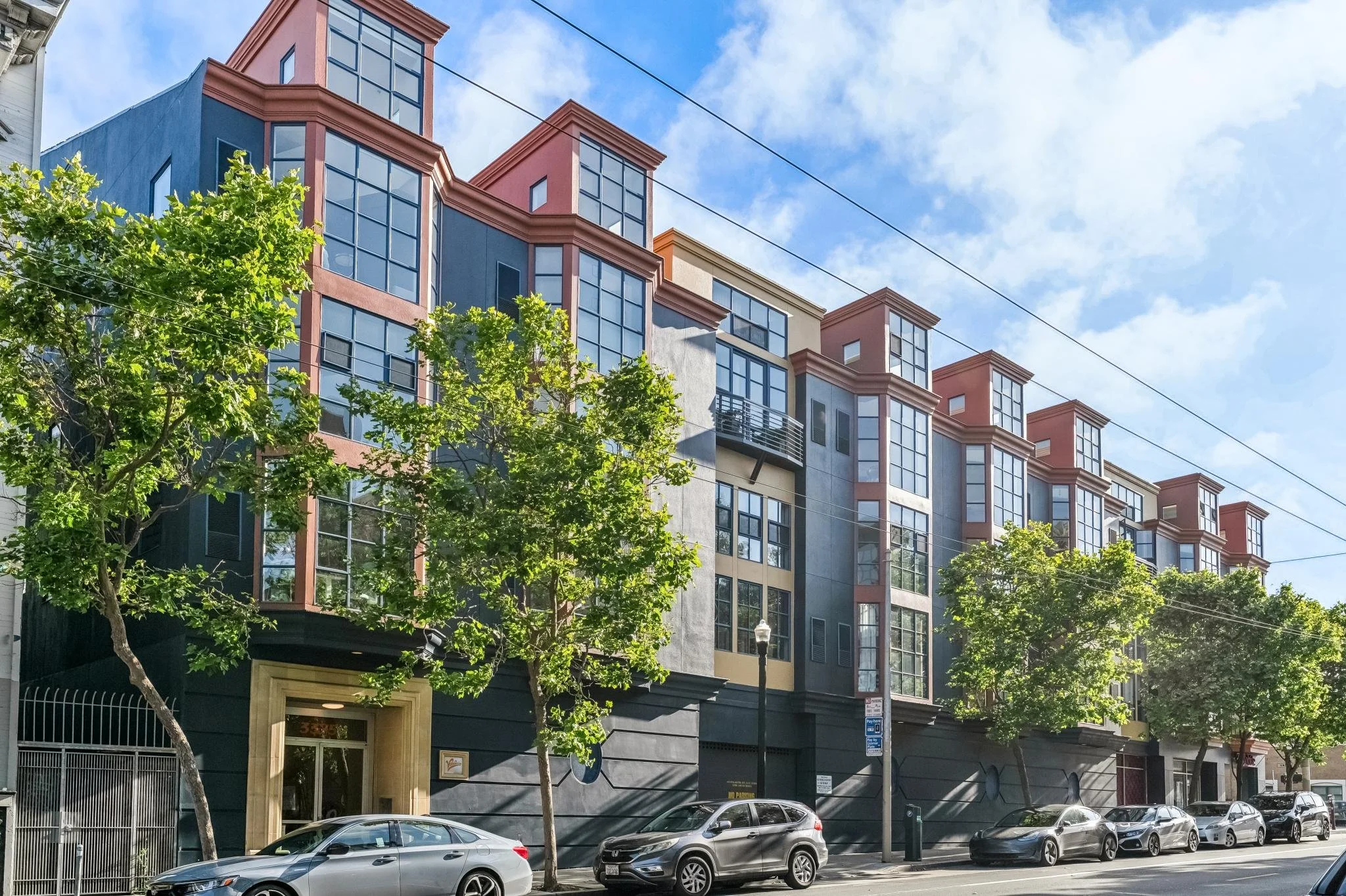 Modern multi-story apartment building with large windows, trees in front, cars parked on street, blue sky with some clouds.