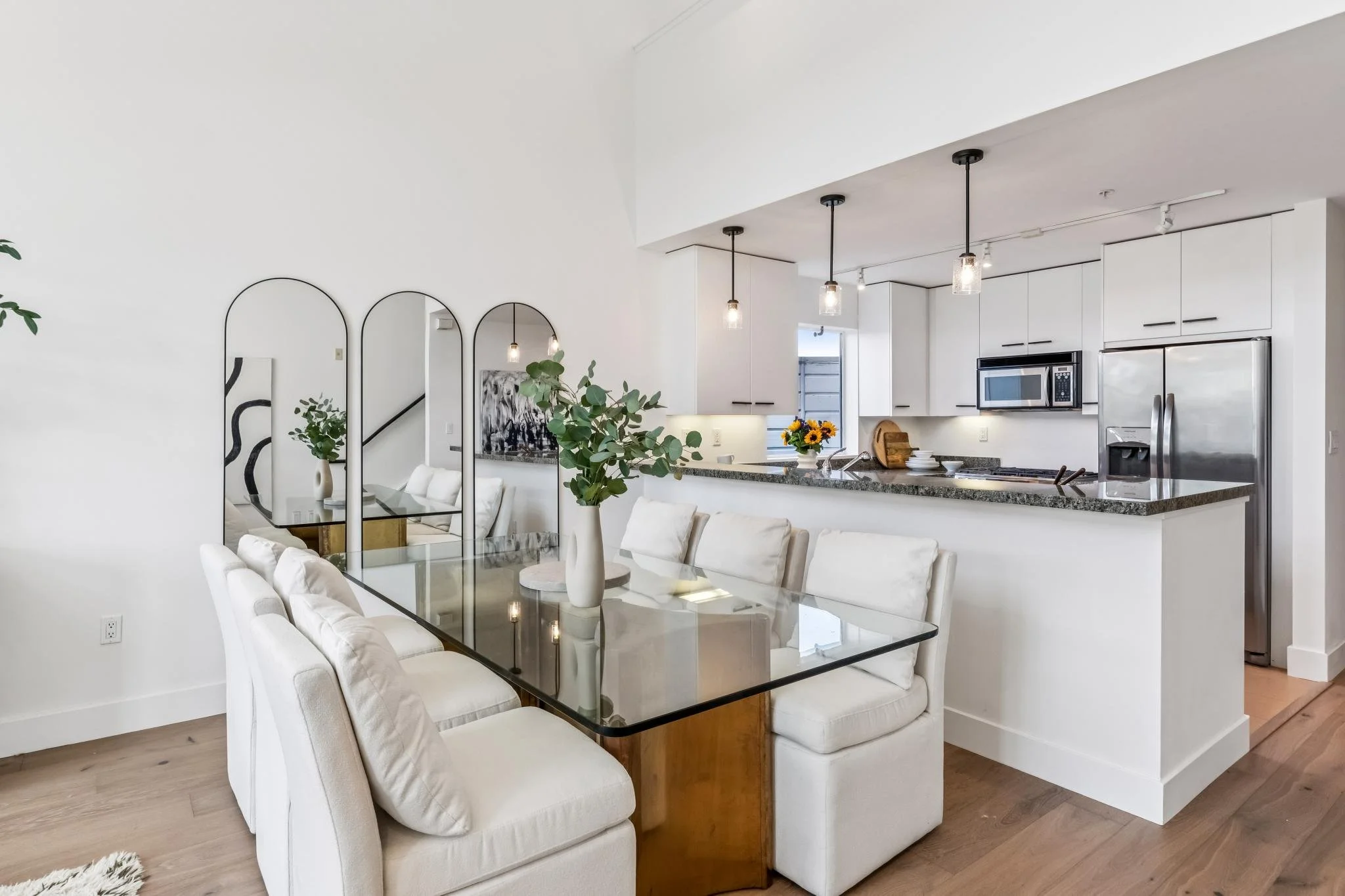 Modern open-concept dining and kitchen area in a bright, white interior with a glass dining table, white upholstered chairs, a kitchen counter with a vase of sunflowers, minimalist decor, and pendant lighting.