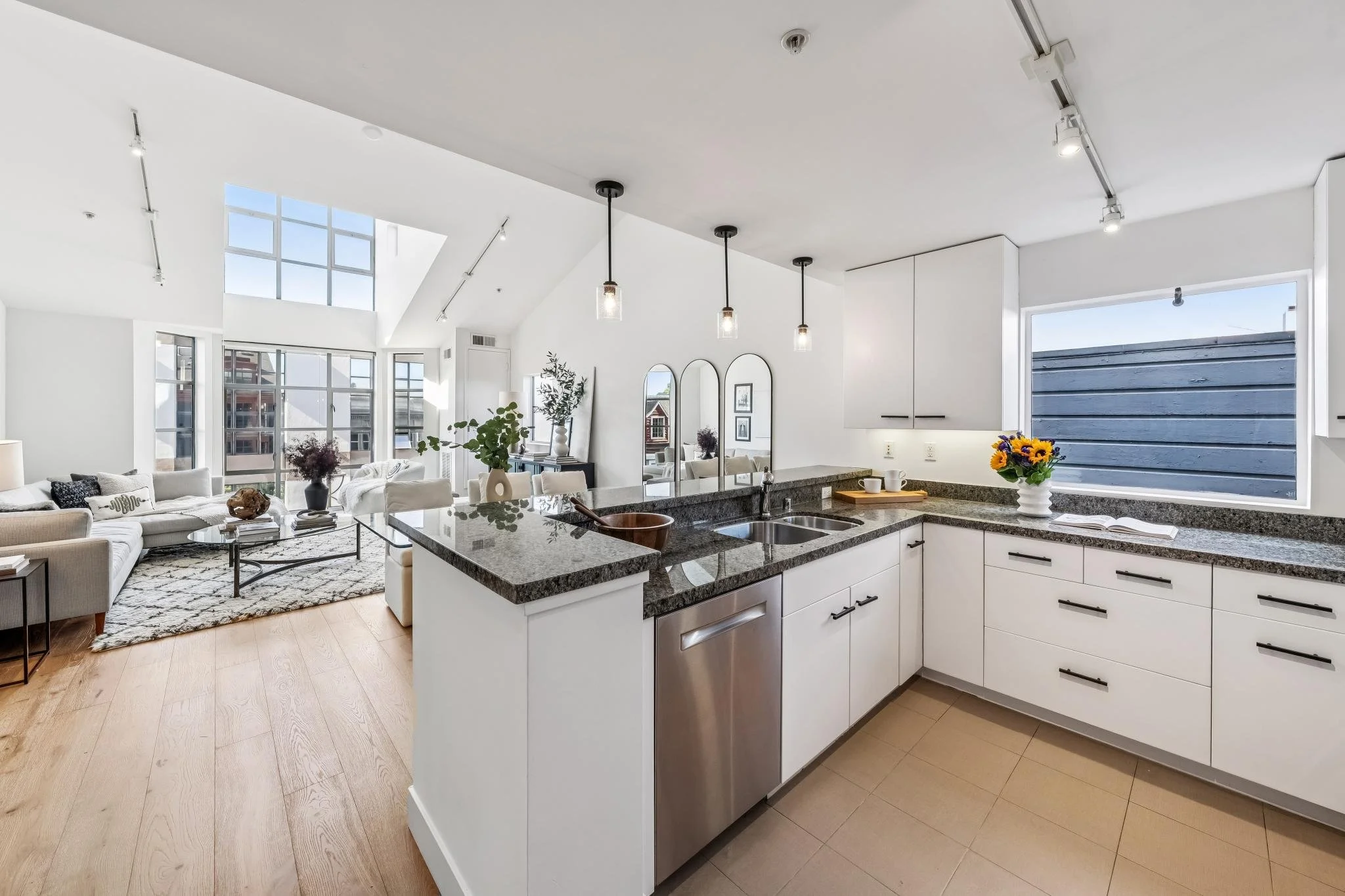 Modern open-concept kitchen with white cabinets, granite countertops, and a breakfast bar, overlooking a bright living room with large windows, plants, and artwork.