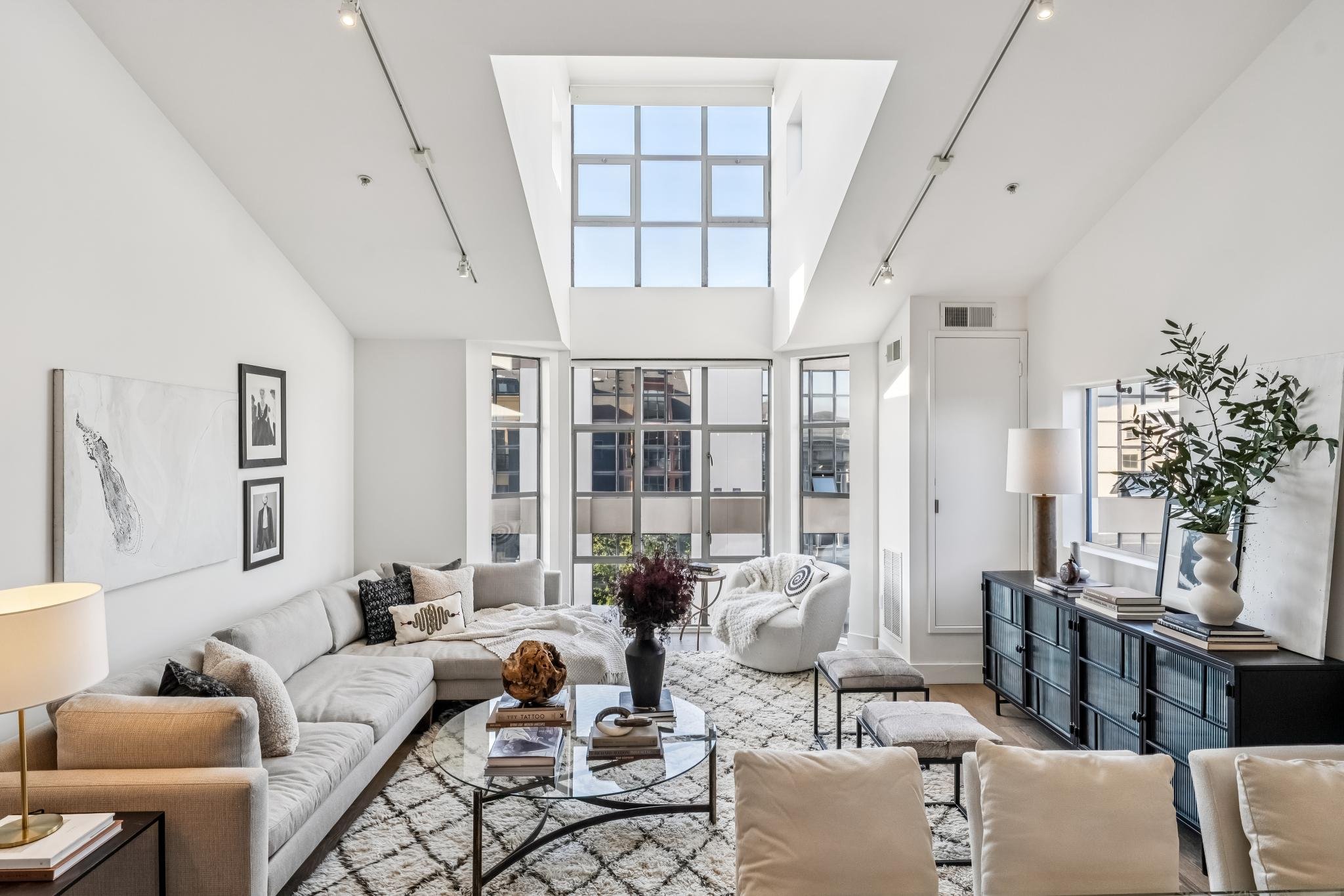 Modern living room with white walls, large windows, beige sectional sofa, black and white artwork, glass coffee table, indoor plants, and natural light from skylight.