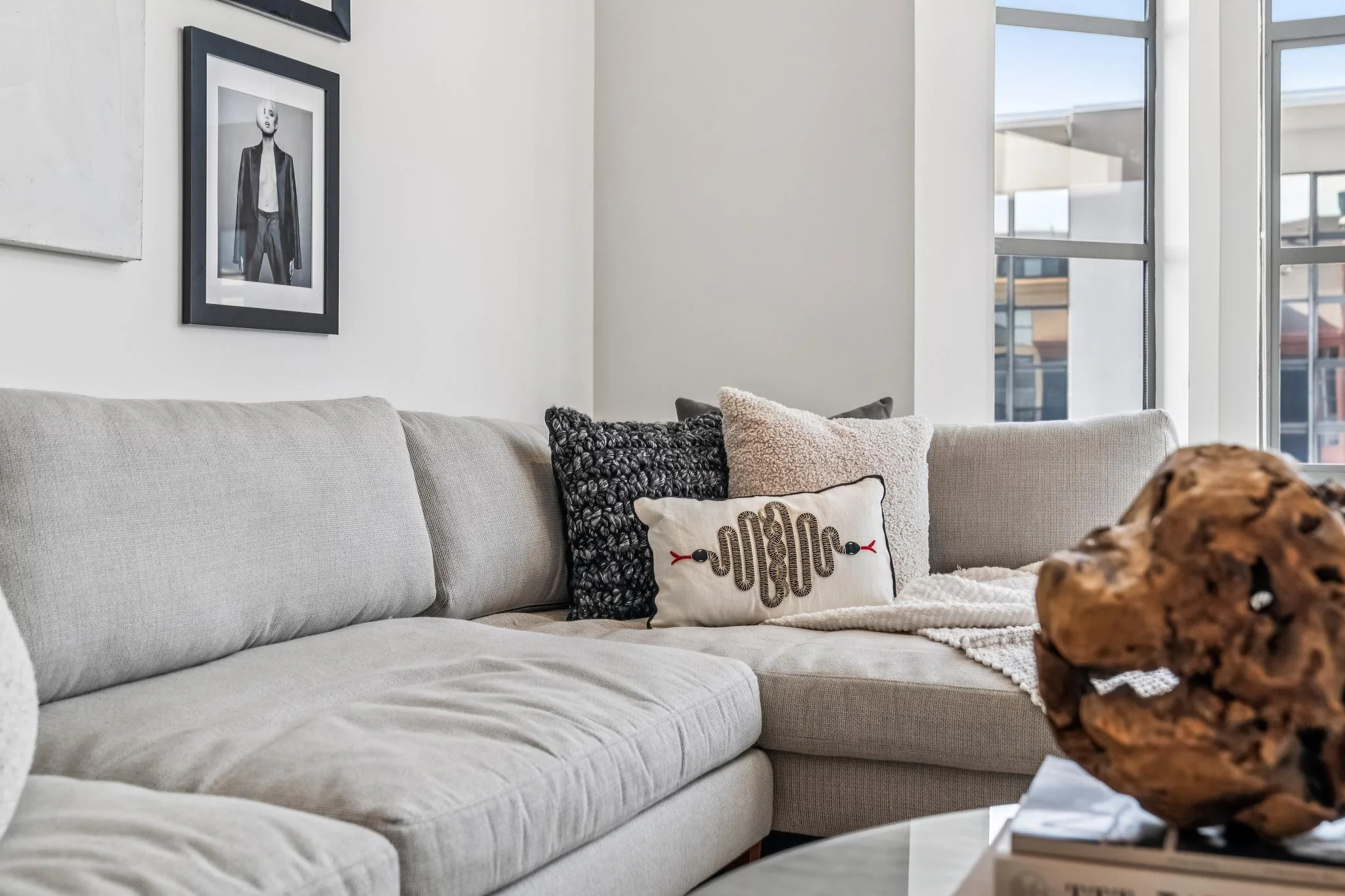 Living room with a light beige sofa, decorative pillows, a glass coffee table with a rocky object, and large windows allowing natural light.