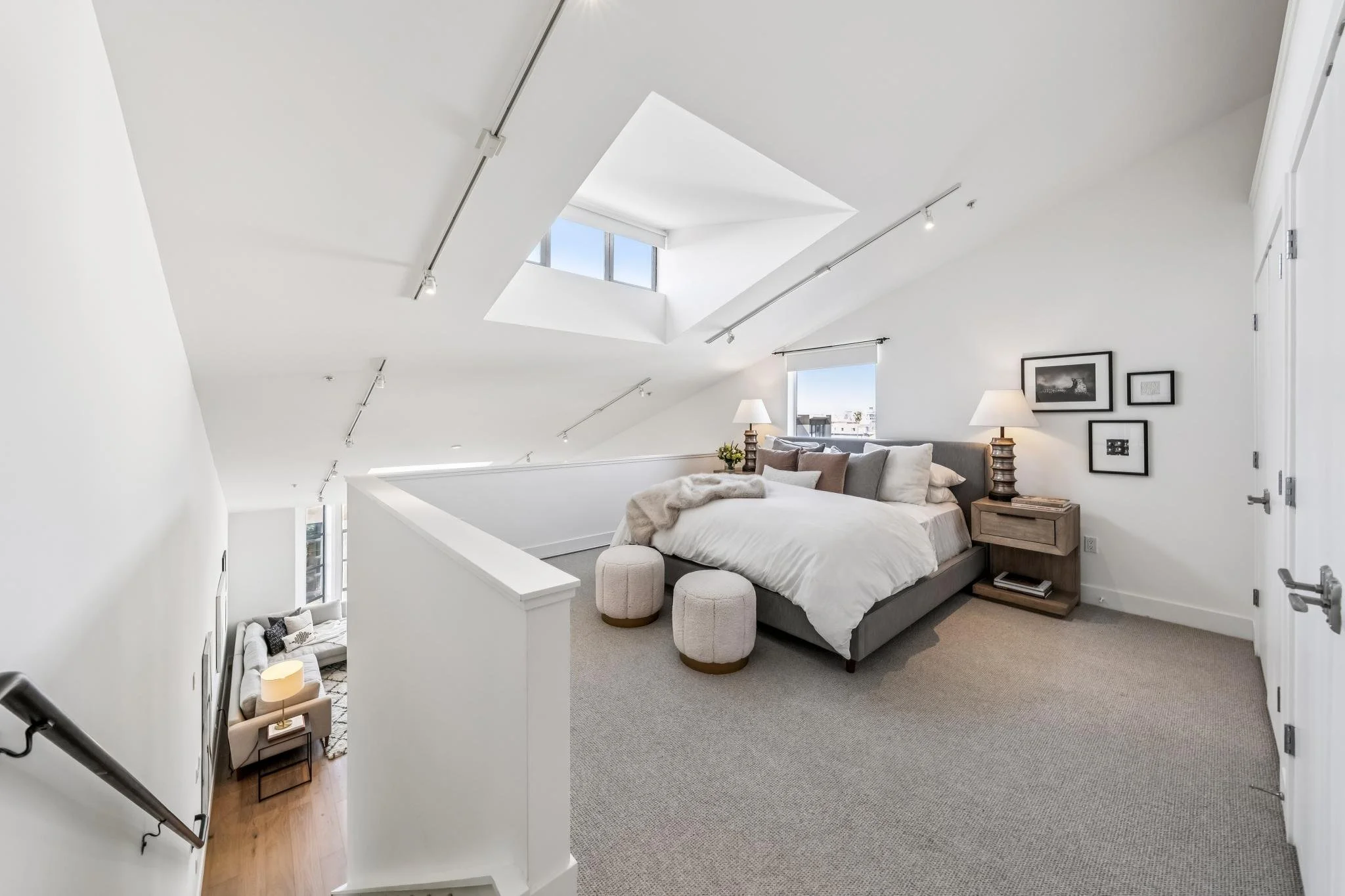 A bright attic bedroom with a sloped ceiling, skylight, and large windows. The room features a bed with white bedding, nightstands with lamps, and framed pictures on the wall. There are two small upholstered ottomans at the foot of the bed.