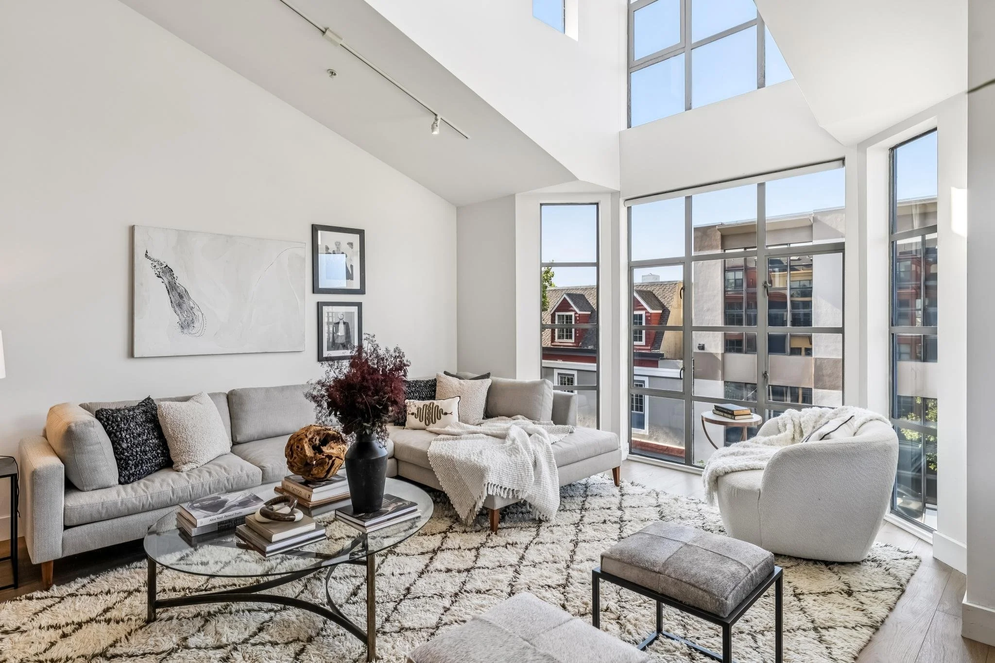 Modern living room with large windows, white walls, beige sofa with black and white pillows, glass coffee table with stacked books and decorative items, white armchair with a blanket, abstract artwork on the wall, and a patterned rug.
