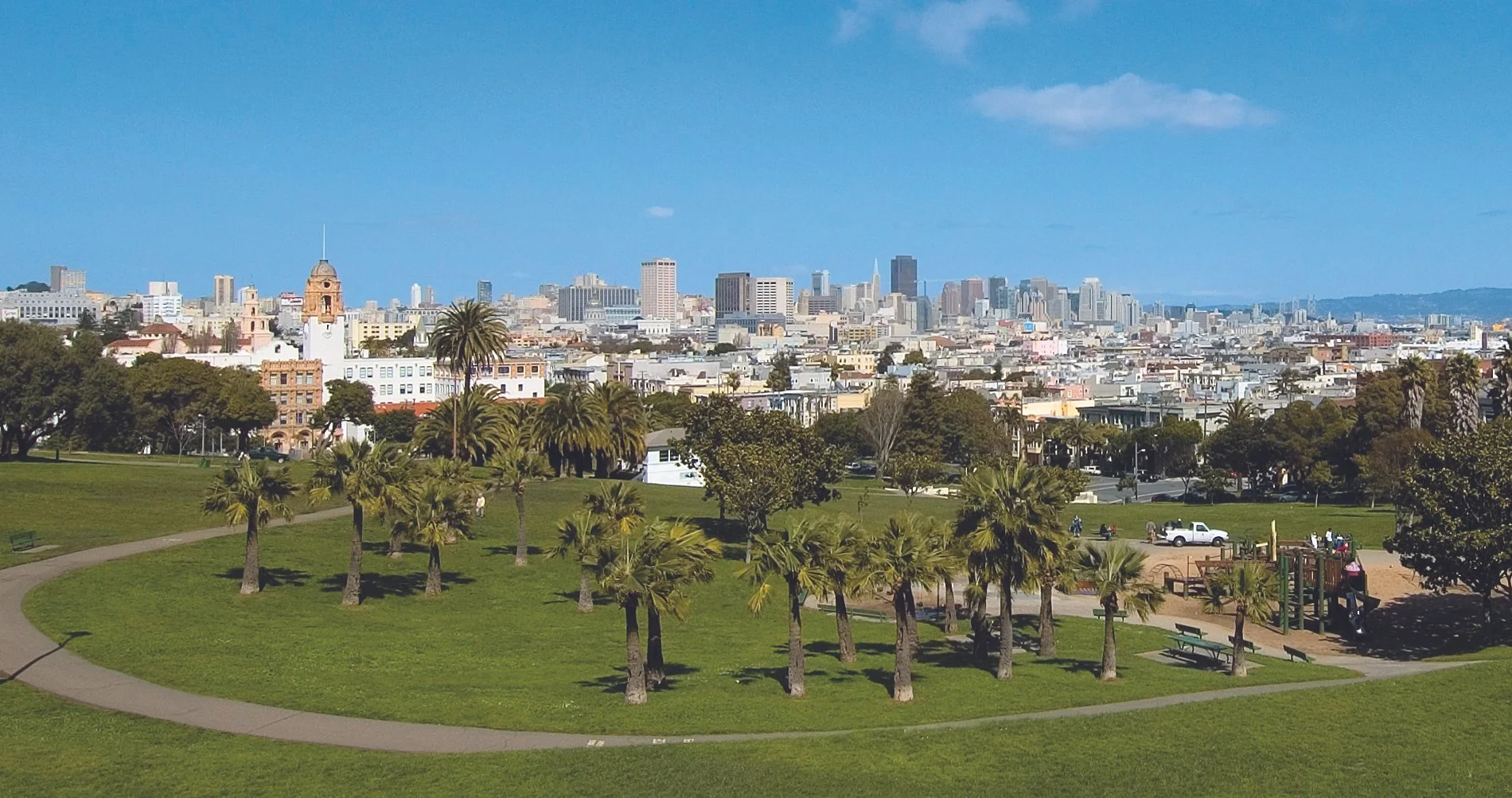 A city skyline view from a park with green grass, palm trees, and a playground in the foreground.