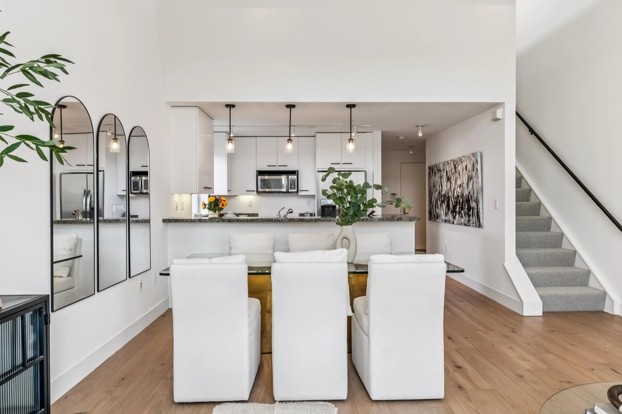 Interior of a modern dining area connected to a kitchen with white cabinets and granite countertops. There are five white upholstered dining chairs around a clear glass table with a decorative vase of flowers. The wall features three industrial-style