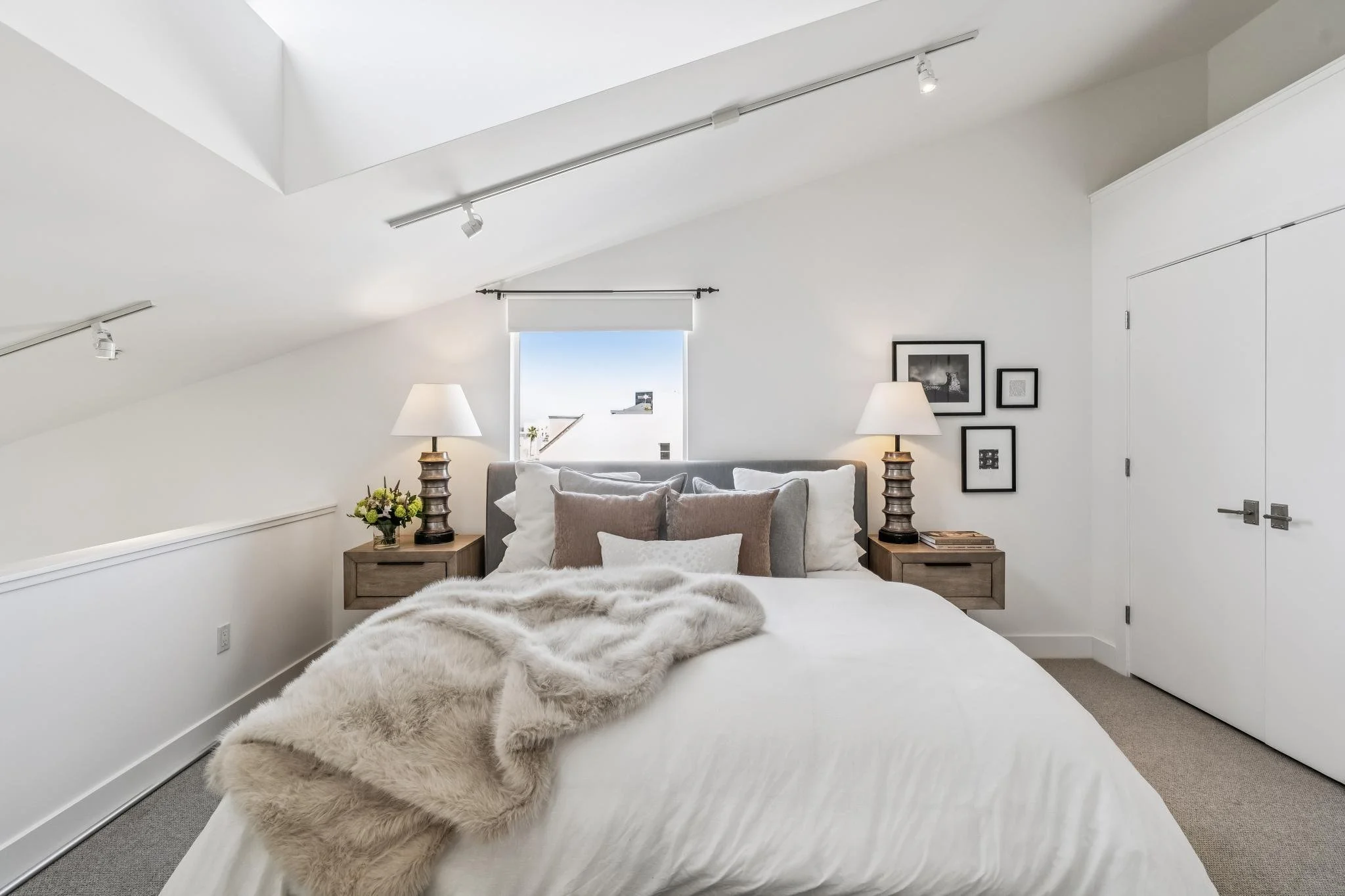 A bedroom with a sloped ceiling, white walls, a window with a white blind, a bed with beige and white pillows, fur throw blanket, and two matching bedside tables with lamps. There are framed pictures on one wall.