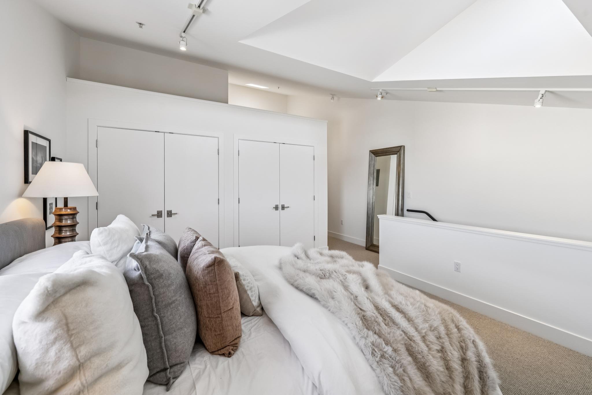 Interior of a modern bedroom with a white color scheme, a white bed with multiple pillows, a lamp on a side table, a large standing mirror, and double closet doors. The ceiling has track lighting and a partial wall separates the sleeping area from th