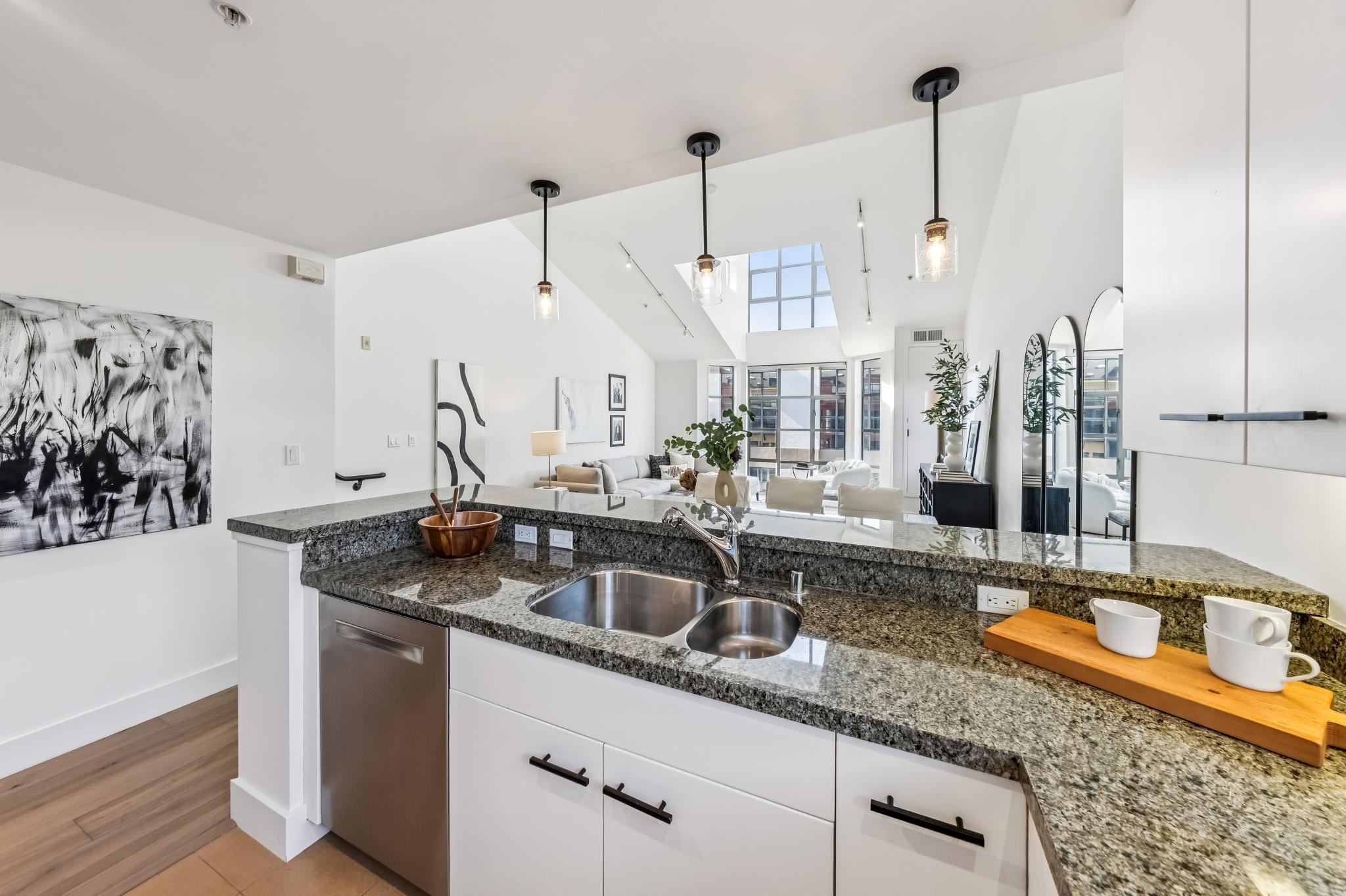 Open-concept modern kitchen with granite countertops, white cabinets, stainless steel sink, and decorative items on the counter, overlooking a bright living room with large windows, artwork, plants, and minimalistic furniture.