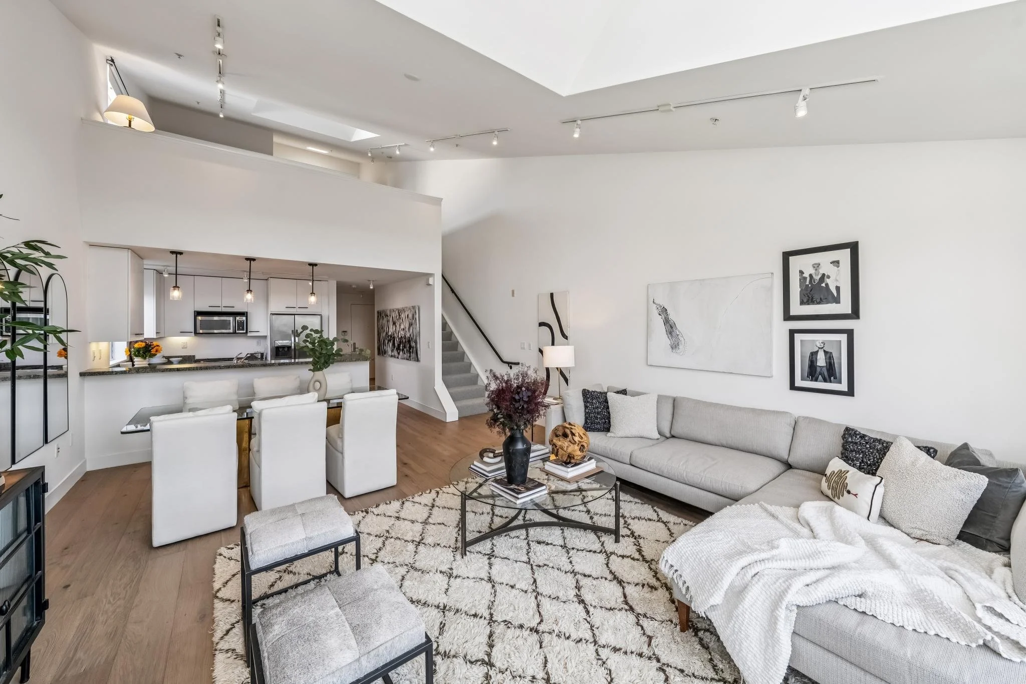 A spacious, modern, open-concept living room with white walls and ceiling, wooden flooring, a large L-shaped sofa with decorative pillows, a glass coffee table with books and decorative objects, framed black and white artwork on the walls, and a view