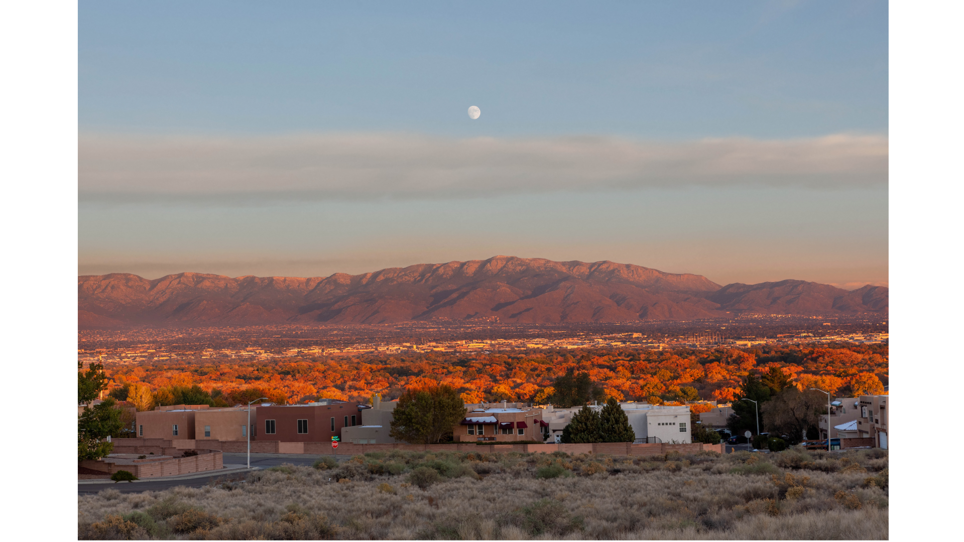 Sunset over a suburban area with mountains in the distance, the moon visible in the sky, and autumn-colored trees in the foreground.