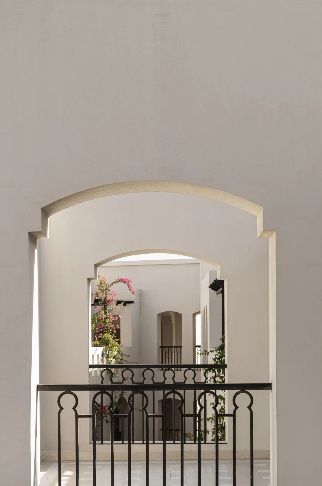 View through a series of archways and windows in a white building with black metal railing and pink flowering plants visible outside.