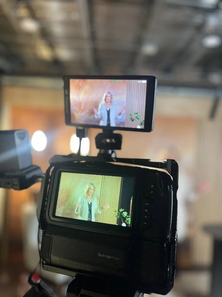 Two cameras capturing a woman speaking on a stage; one camera's screen shows her in front of a colorful background, and the other camera's screen shows her in front of a green backdrop with vertical lines.