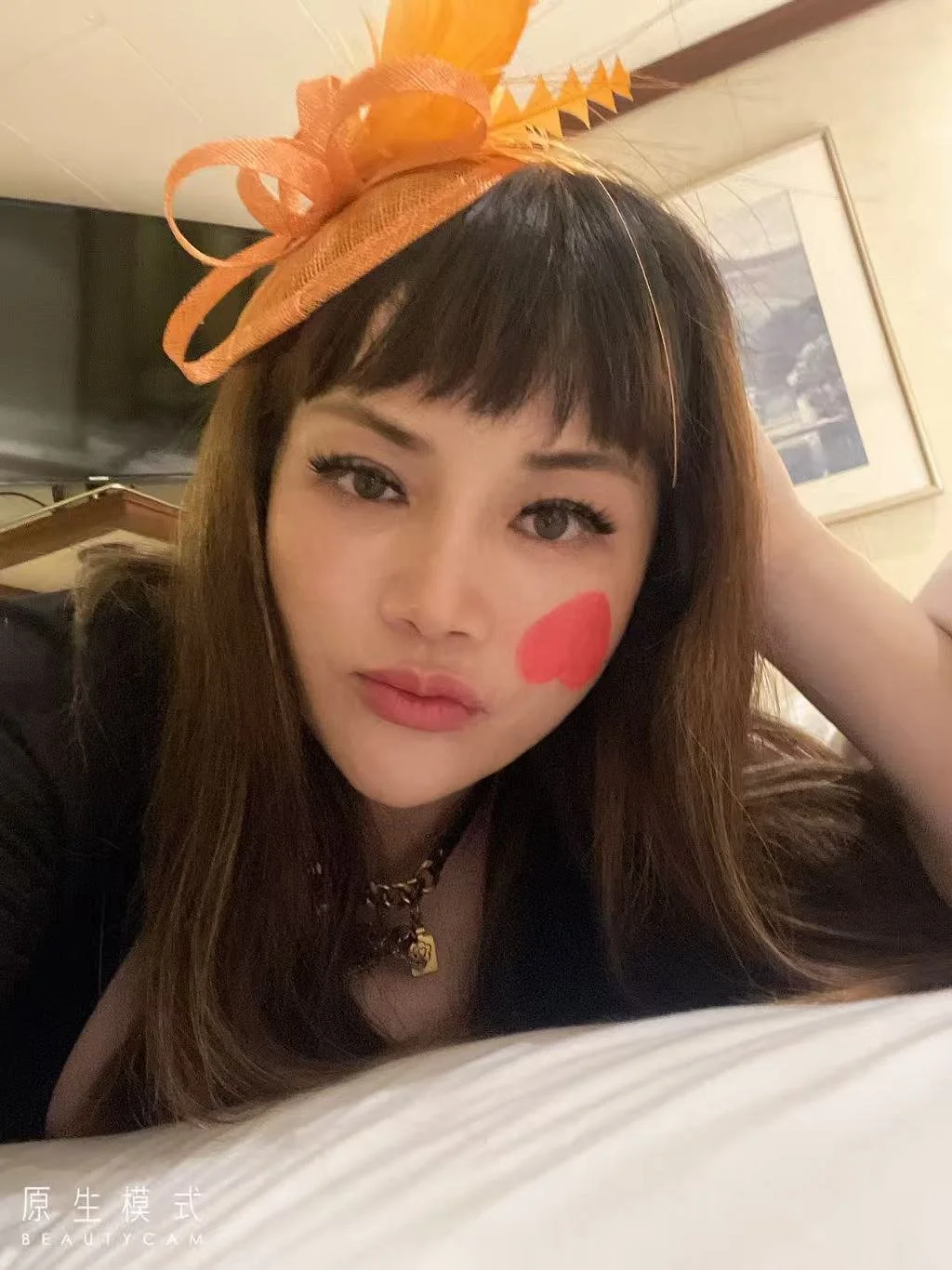 A woman with brown hair and bangs wearing an orange hat with a bow, lying down with her head resting on her hand. She has a red heart painted on her cheek and is wearing makeup with eyeliner and lipstick. She is dressed in black with a gold necklace and is in a room with a framed picture on the wall behind her.